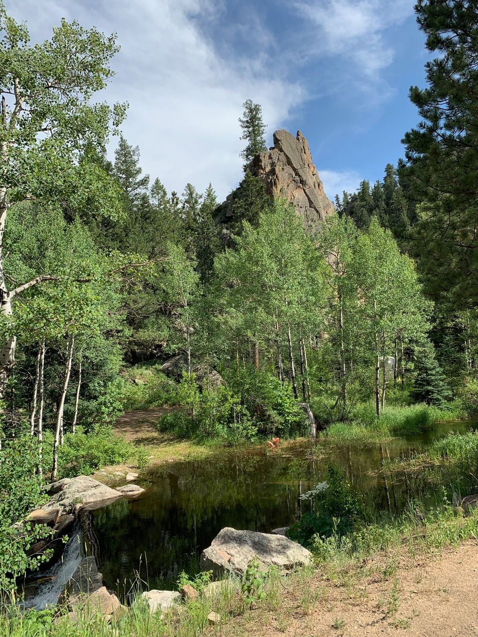 Rocky mountain peak rises above lush green trees, with a small stream in the foreground under a cloudy blue sky.