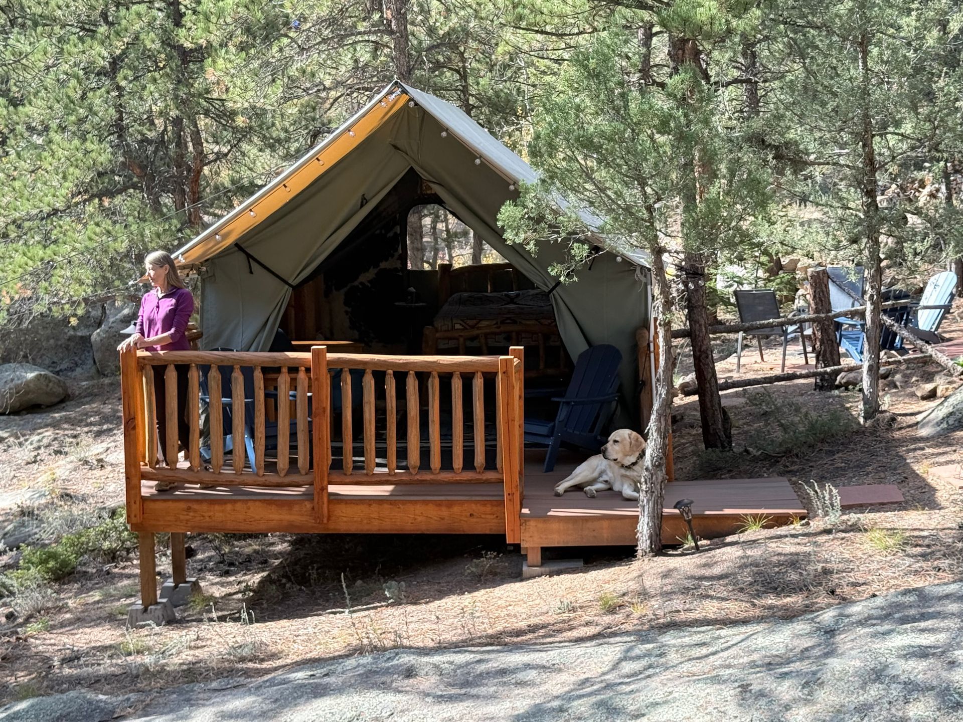 Woman stands on a wooden deck with a dog near a canvas tent in a wooded area.