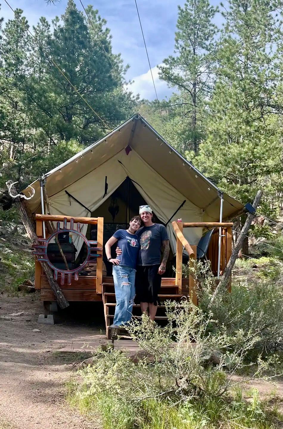 Couple standing on the porch of a canvas tent in a wooded area, smiling.