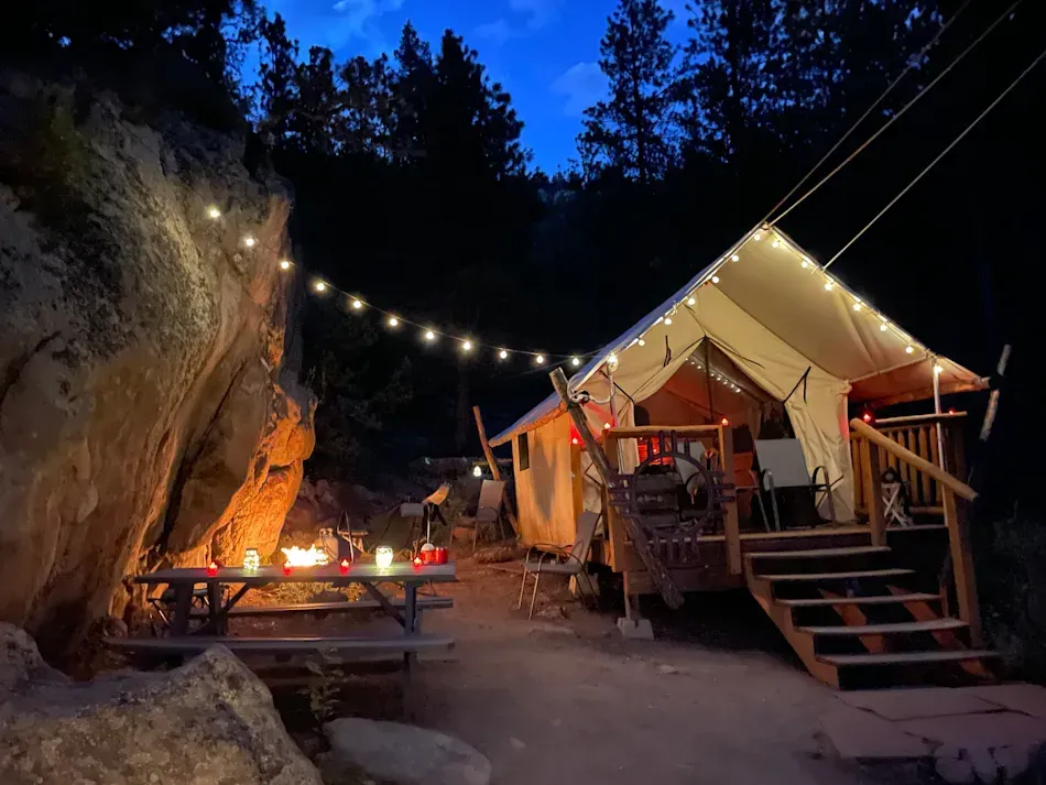 A lit canvas tent with a wooden deck at dusk, beside a rock face, string lights overhead.