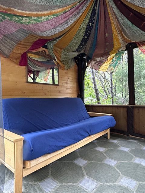 Cozy screened porch with blue futon, decorated fabric ceiling, wooden walls, and a view of trees.