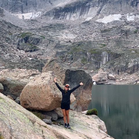 Person hikes on a path towards mountains with a backpack. Green hillside, trees, and gray mountains.