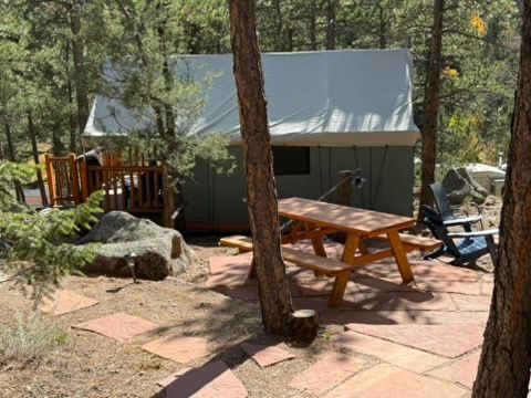 Glamping setup with tent, picnic table, and chairs on a stone patio in a wooded area.