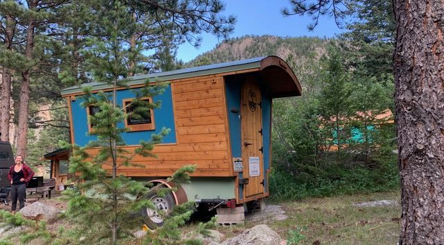 A small, wooden cabin on wheels, painted blue and brown, is parked in a wooded area with a mountain in the background.