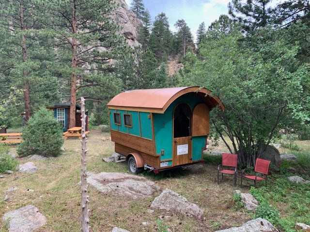 A teal and wood-colored gypsy wagon on a trailer, parked near trees and rocks; two red chairs sit outside.