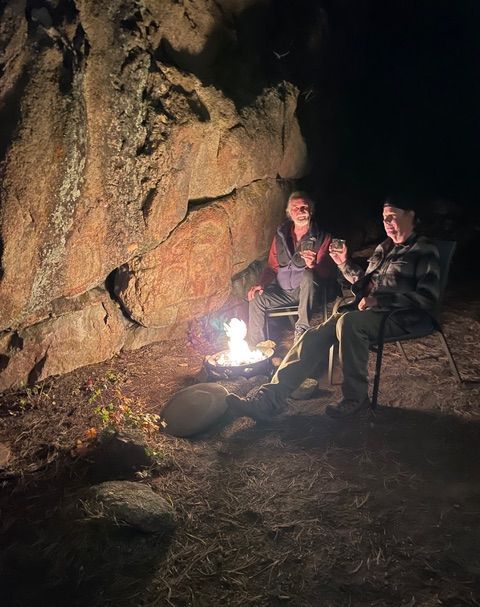 Two people toast by a campfire near a large rock at night.