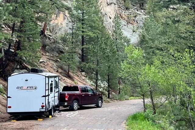 Red pickup truck towing a white camper on a dirt road in a forested area.