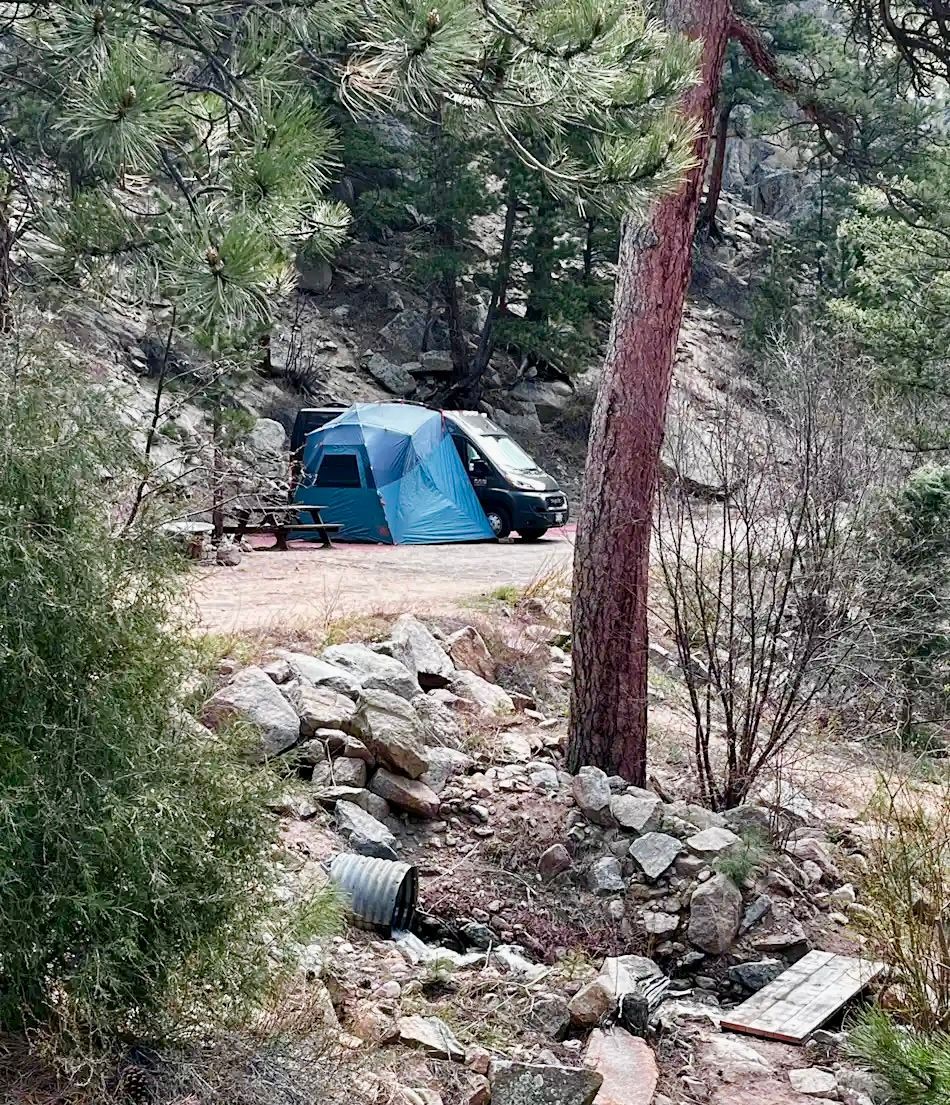 Blue tent and car at a campsite in a forested area, with a stream and stone wall in foreground.
