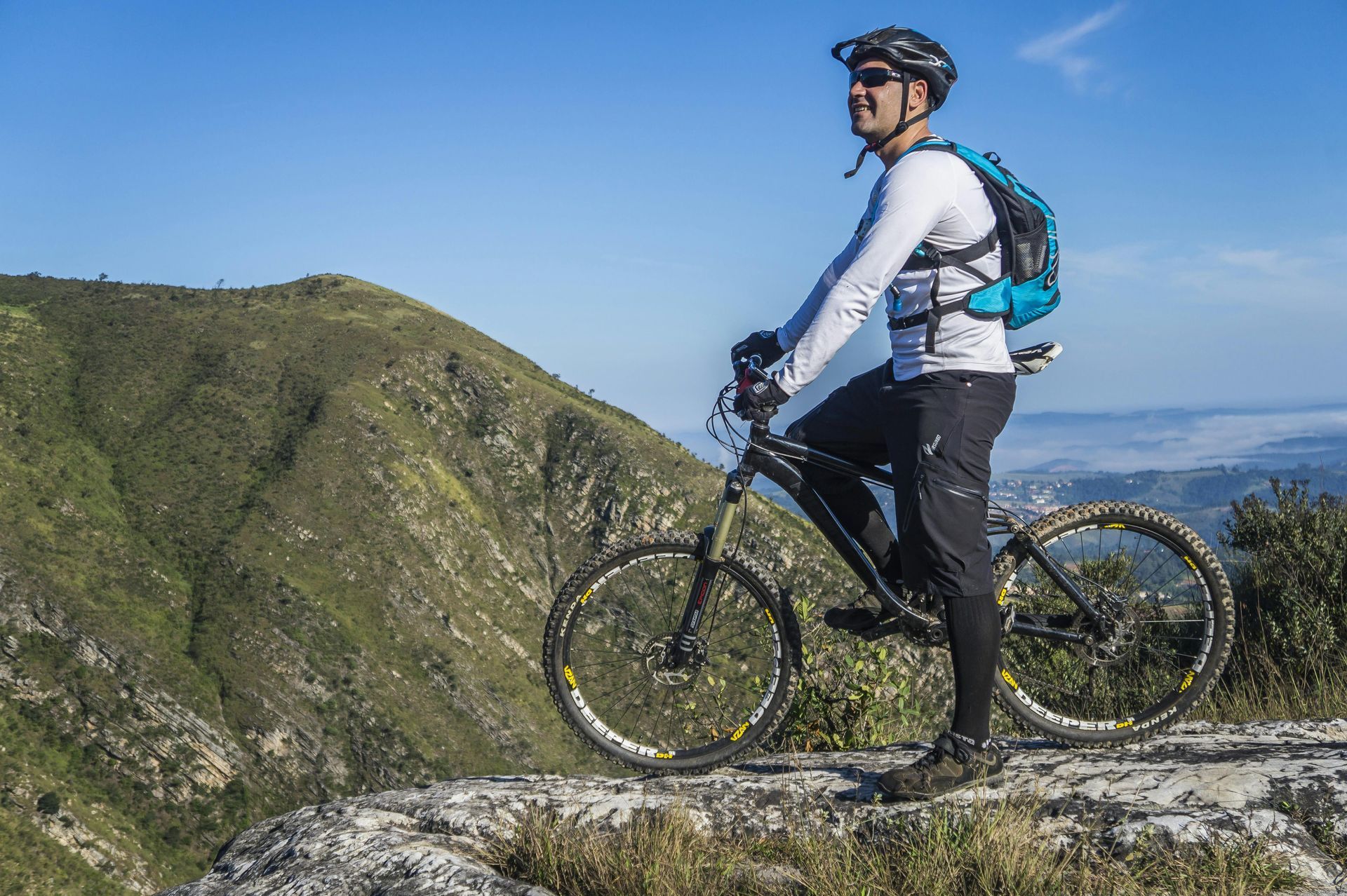 Cyclist on a mountain bike overlooking a green mountain range under a blue sky.