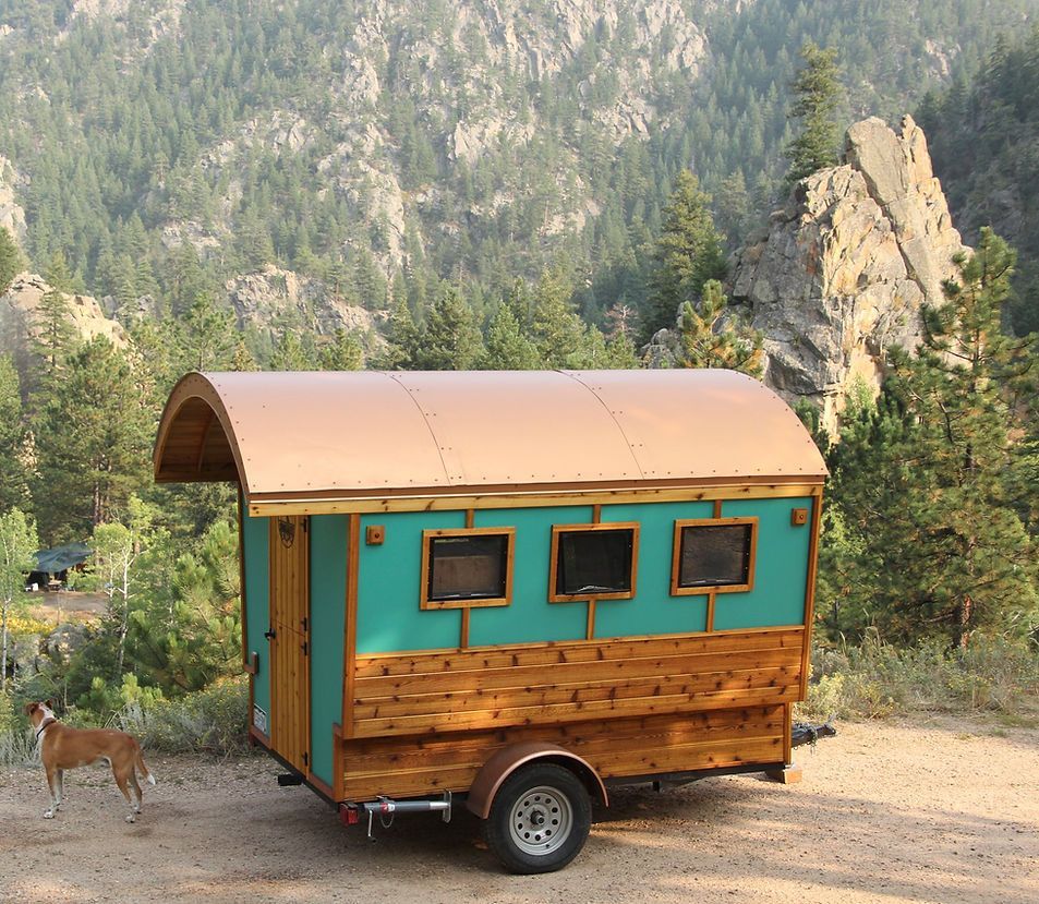 Tiny, teal and wood-paneled camper on a trailer, with a brown roof, parked near a rocky mountain with a dog.