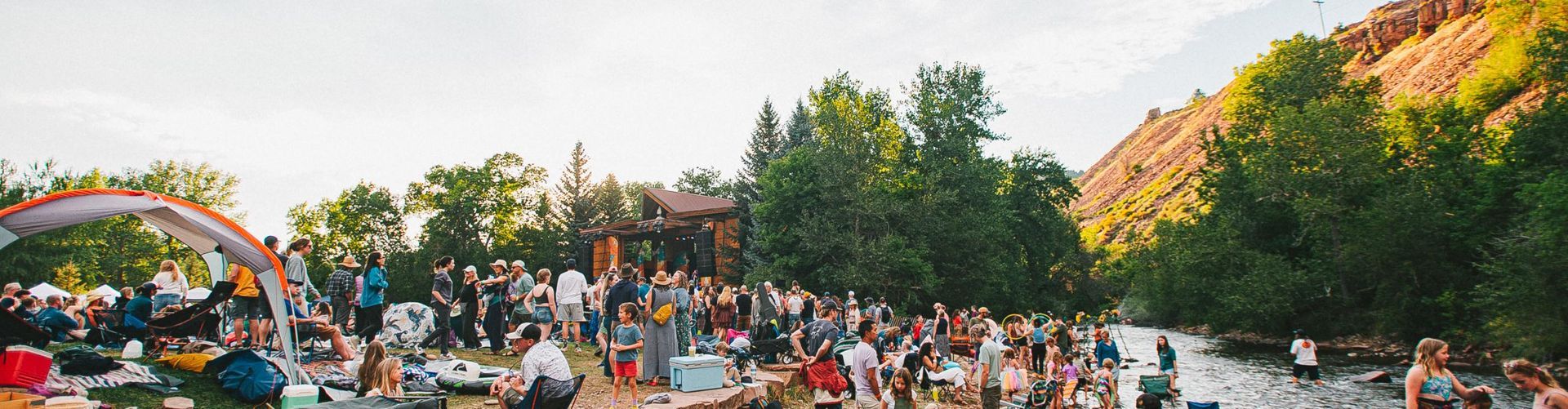 Large crowd gathered outdoors near a river and trees, likely a celebration or event with a mountain in the background.