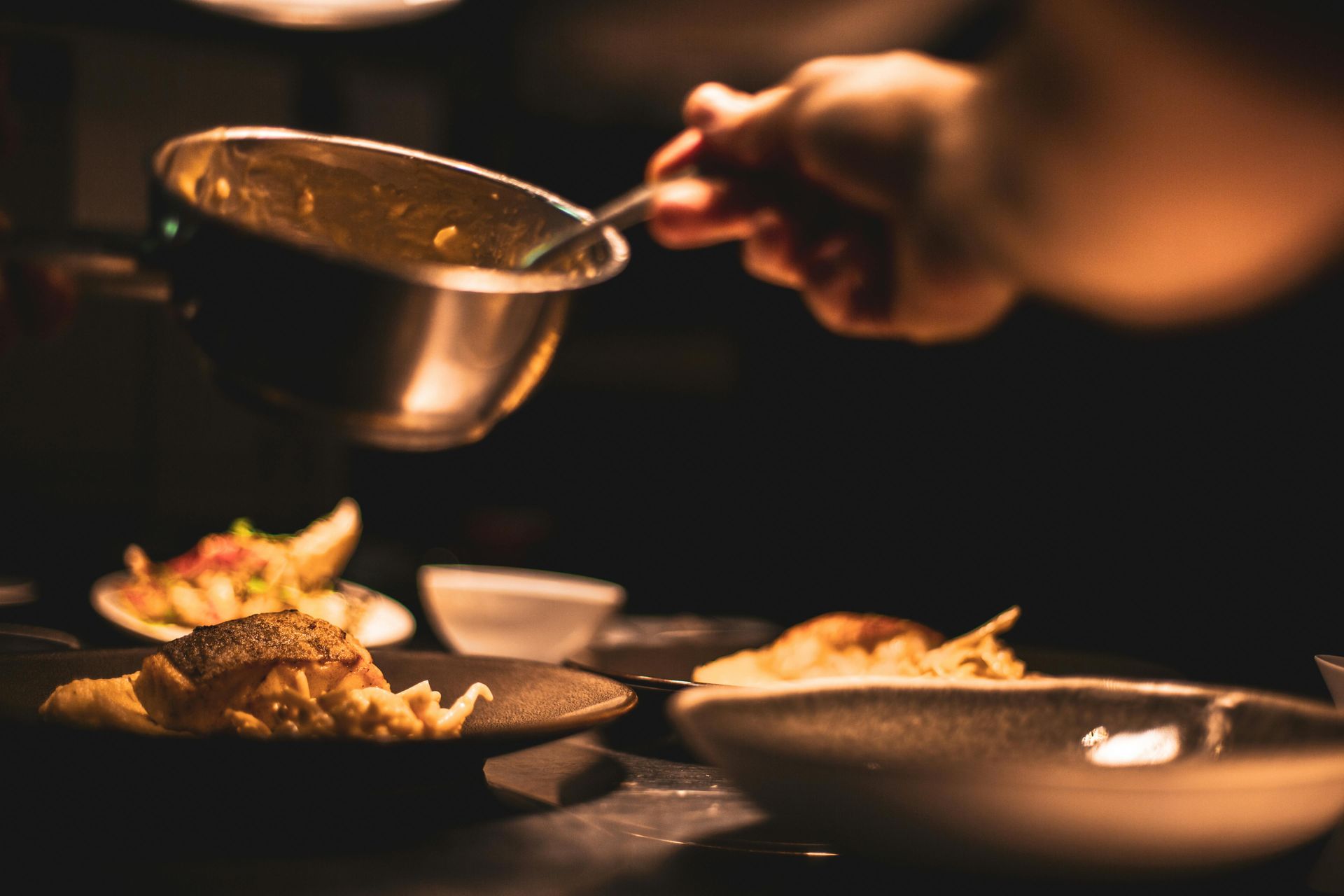 Chef pouring sauce from a silver saucepan onto a plate of food in a dimly lit kitchen.