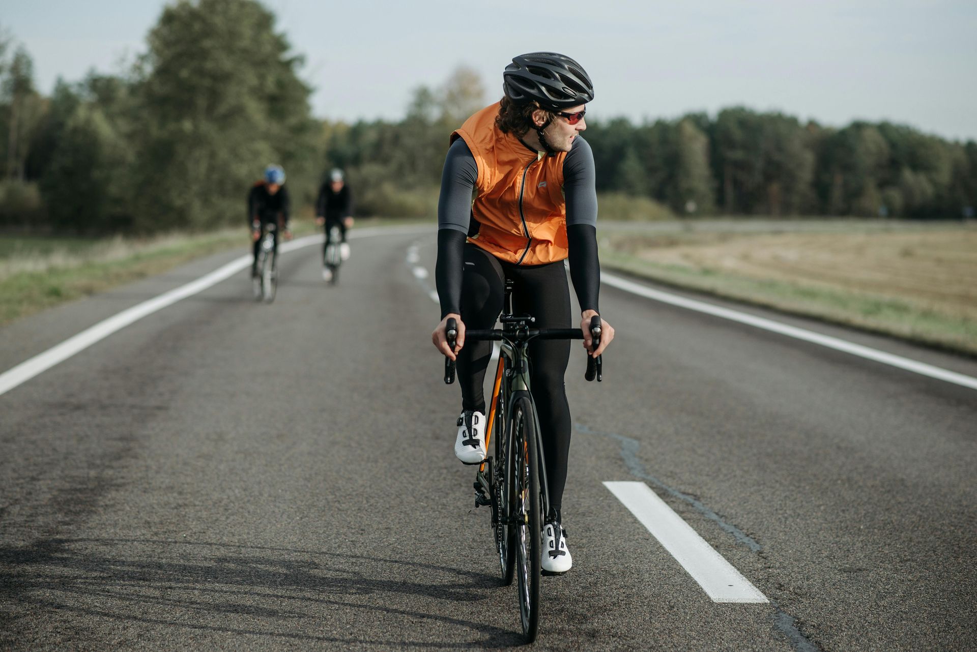 Cyclist in orange vest rides on road with two other cyclists in the background.