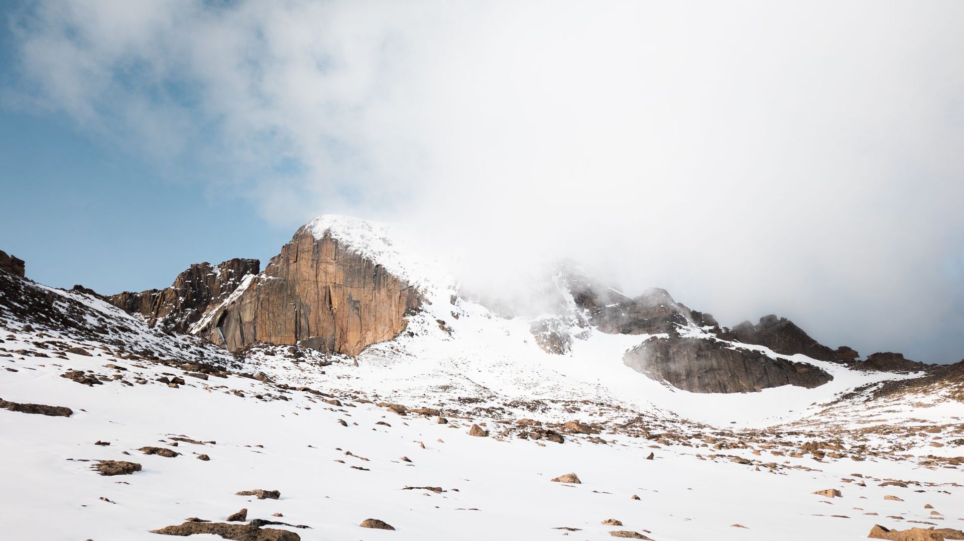Snow-covered mountain peaks with rocky cliffs, partially obscured by clouds, under a blue sky.