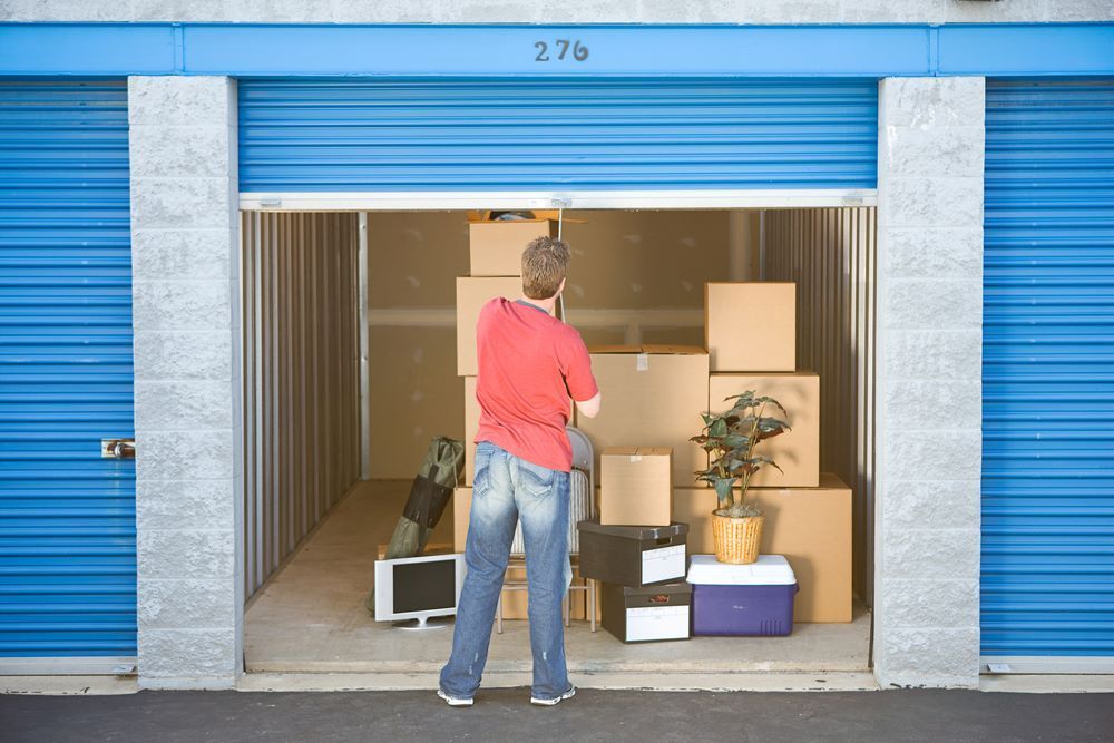 A Man in a Red Shirt is Standing in a Storage Unit Filled With Boxes — AKM Self Storage In Kurri Kurri, NSW
