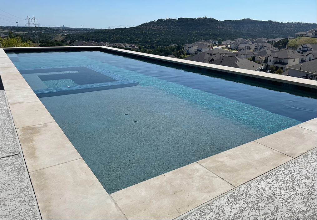 Rectangular swimming pool with light gray tile and water, with a view of houses and hills in the background.