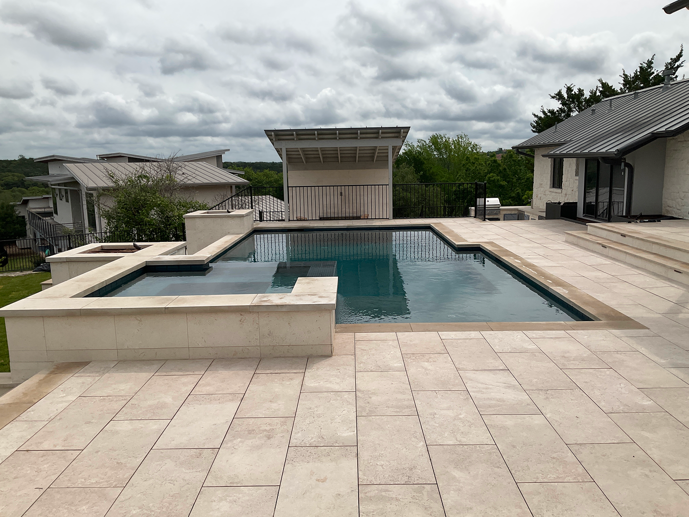 Swimming pool with a spa, on a stone patio, with a cabana and house in the background. Overcast sky.