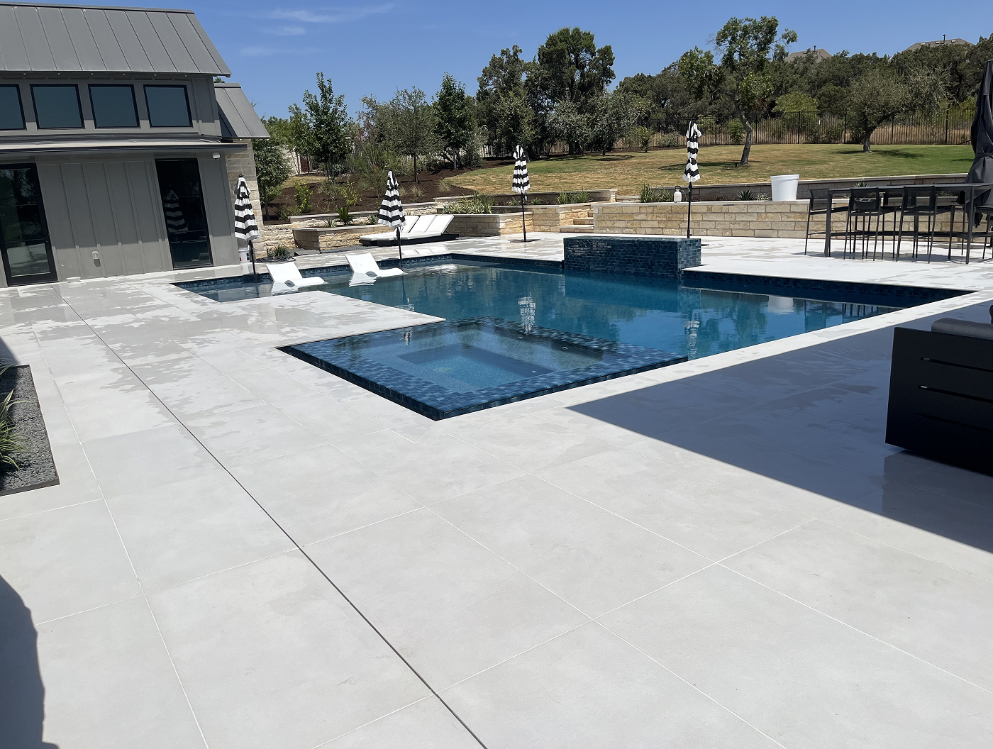Pool area with blue tiled pool, concrete patio, modern house, and lounge chairs on a sunny day.