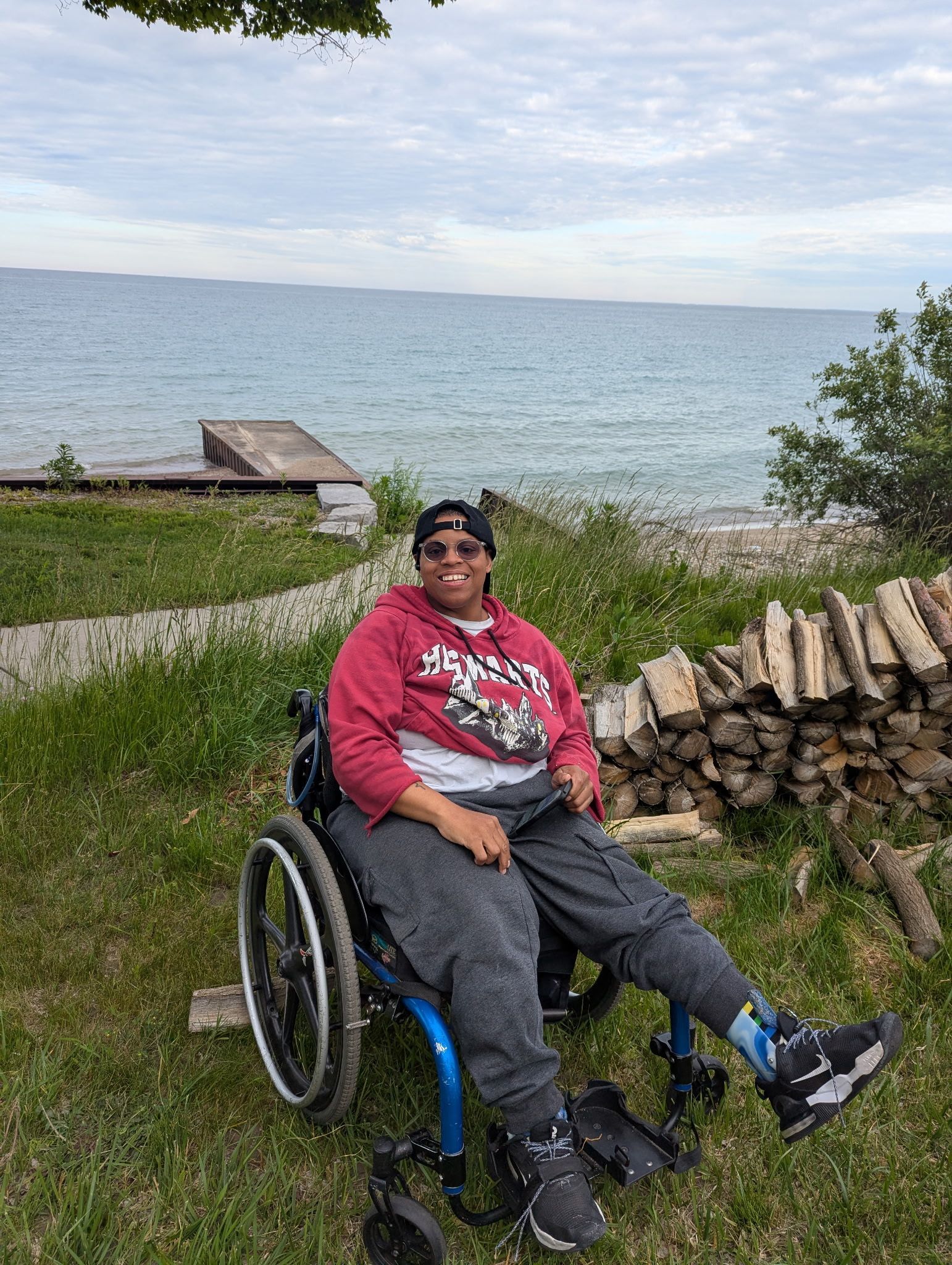 Person in wheelchair smiles by a lake, wearing red hoodie, gray sweatpants, and black cap.
