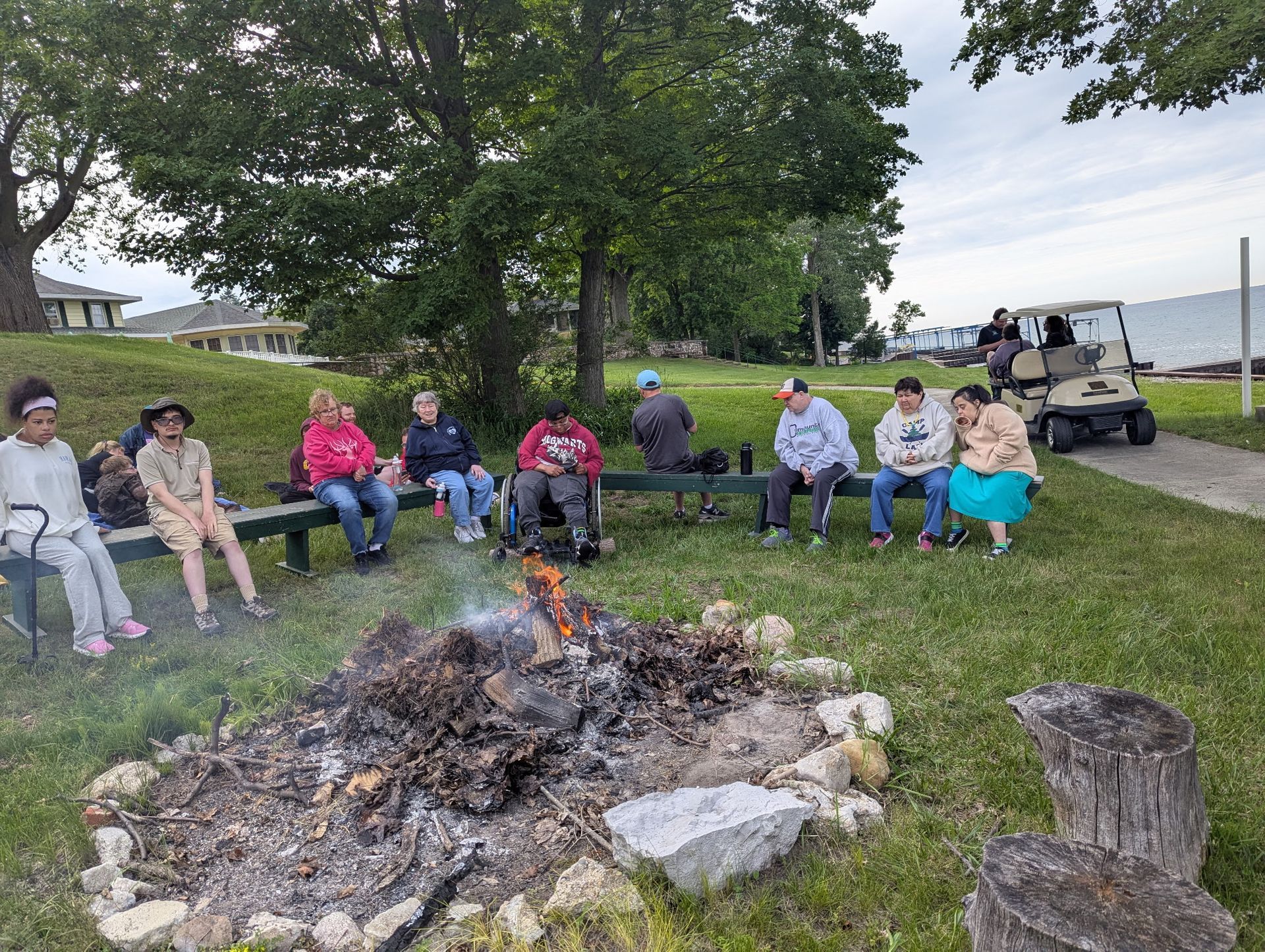 People sitting around a campfire on a grassy lawn near a lake. A golf cart is parked nearby.