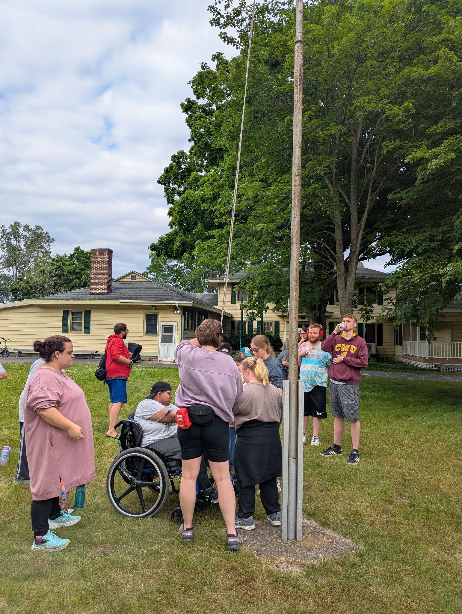 Group of people around a tall pole, some reaching up. Green grass, houses, overcast sky.