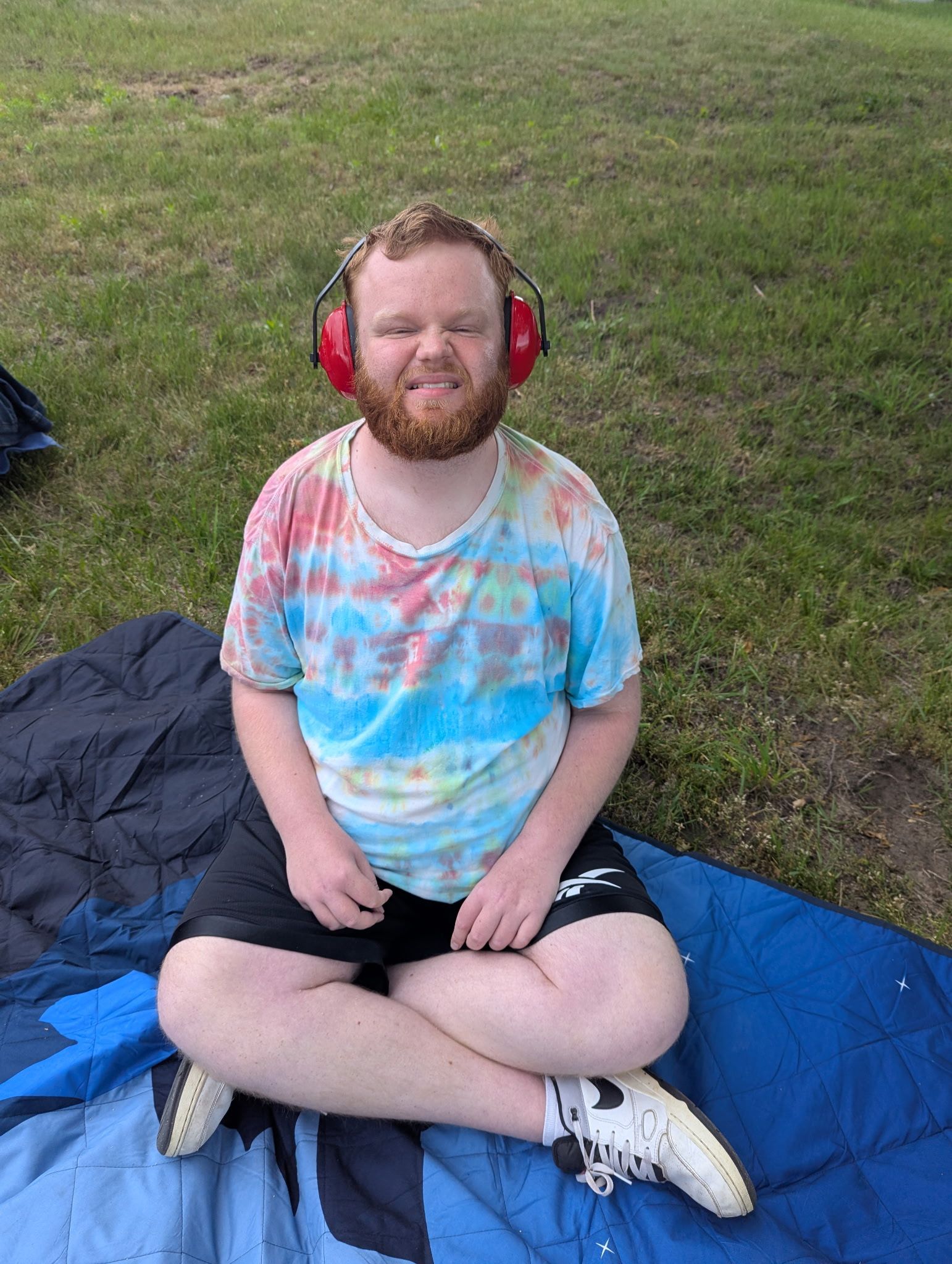 Man with a red beard wearing earmuffs and a tie-dye shirt, sitting cross-legged on a blanket outdoors.