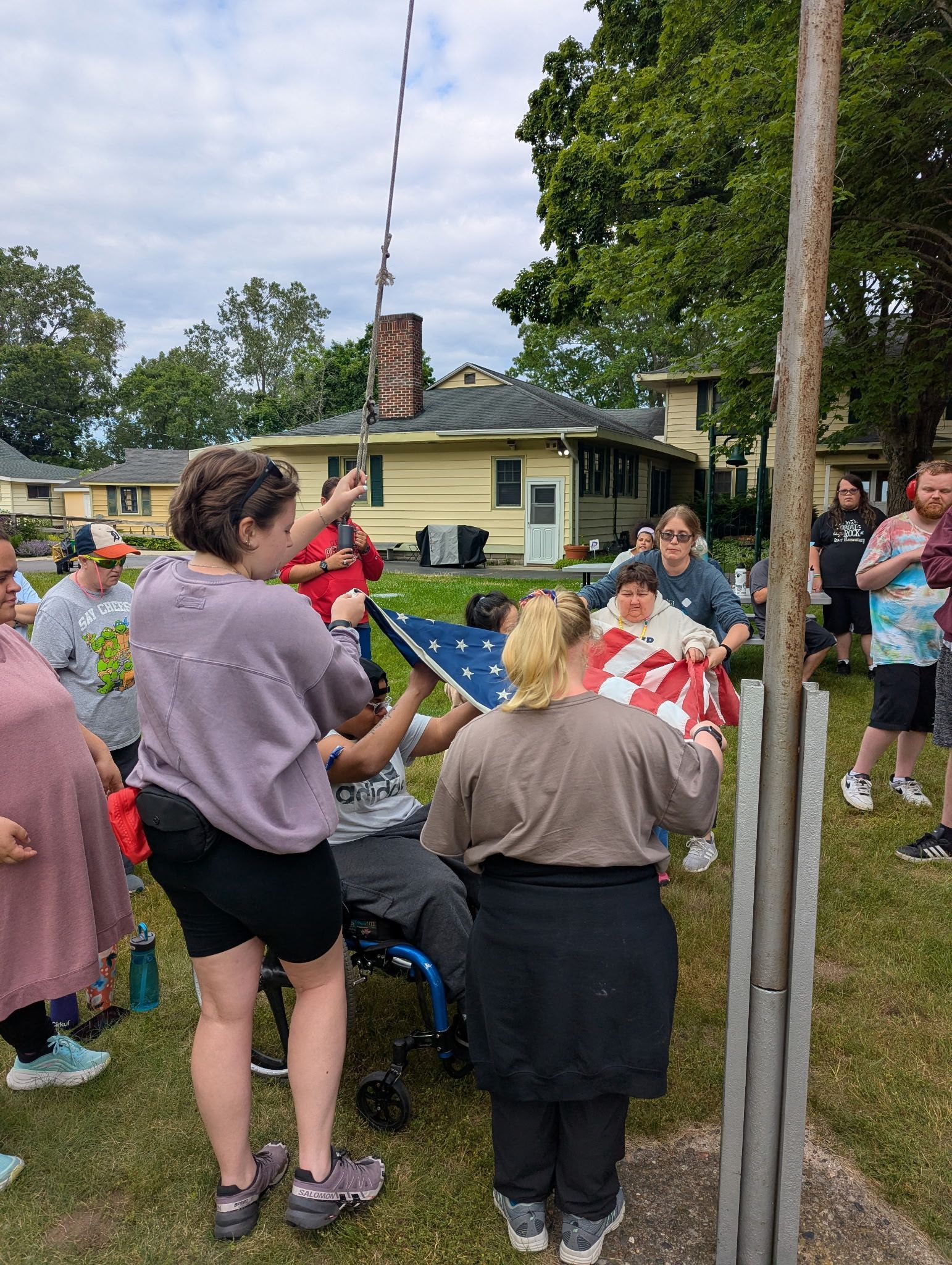Group of people raising the American flag outdoors. People assist from a wheelchair as others watch.
