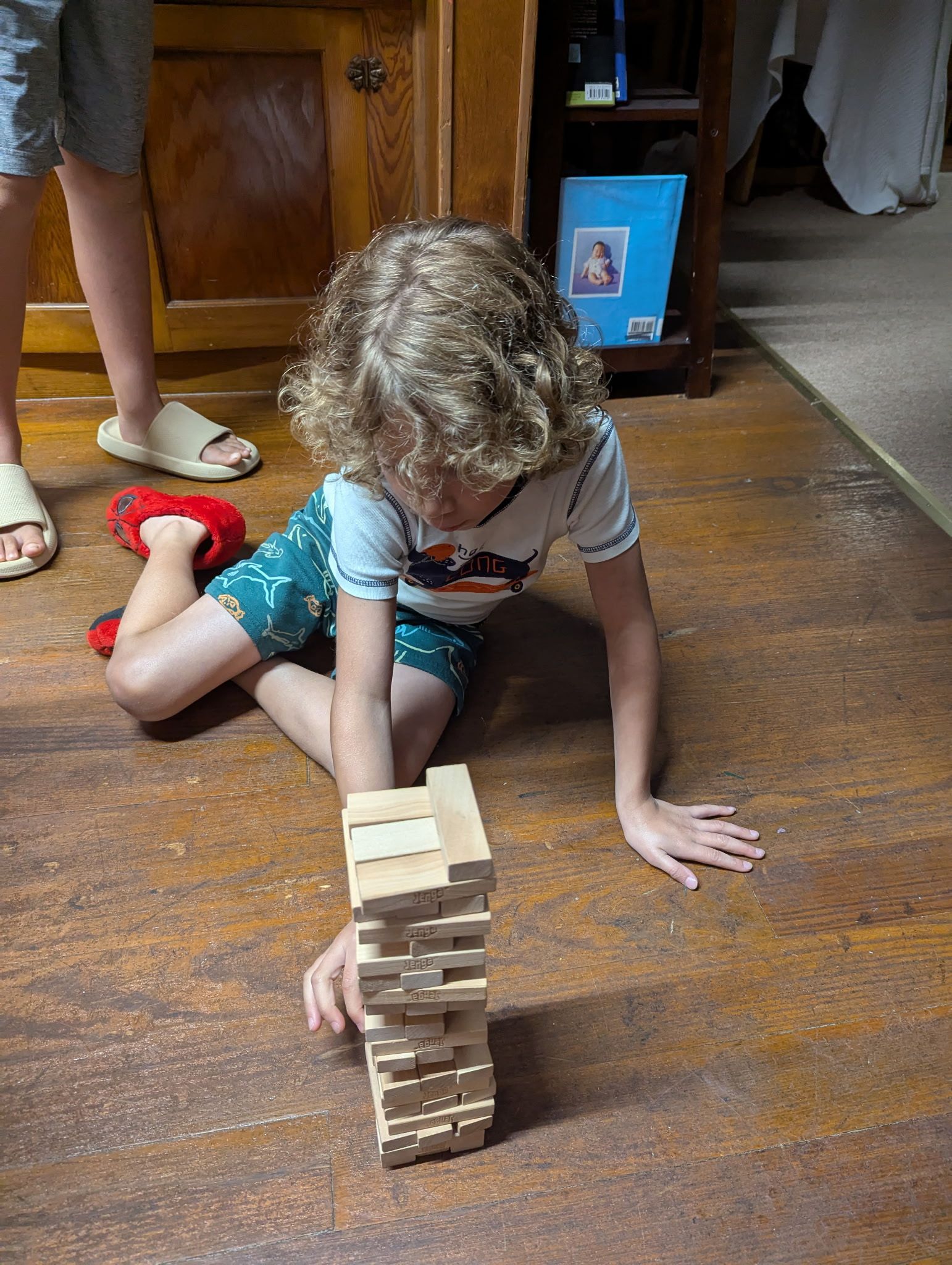 Child playing Jenga on a wooden floor, reaching for a block. Another person's feet in background.