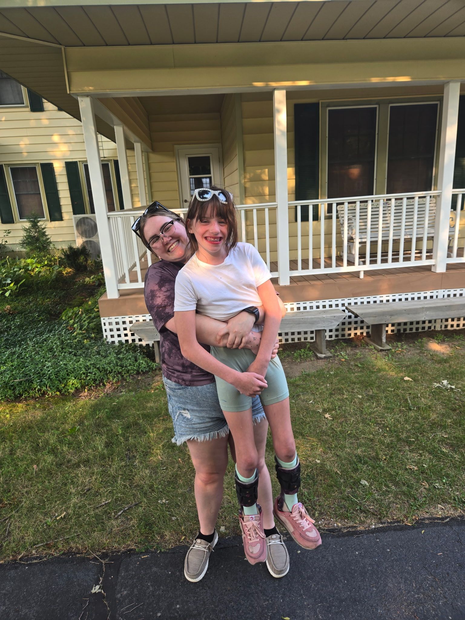 Woman holding girl, both smiling in front of a yellow house. The girl wears leg braces and pink shoes.