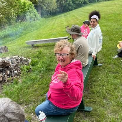 A group of people by a campfire on a grassy hill. A woman in pink points at the viewer, smiling.