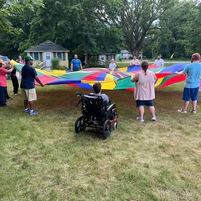 People playing with a colorful parachute in a grassy yard; person in wheelchair is centered.