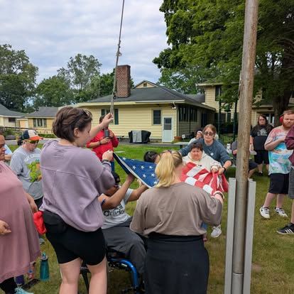 People raising an American flag outdoors. Several people hold the flag while others work the rope. Green grass, yellow houses.