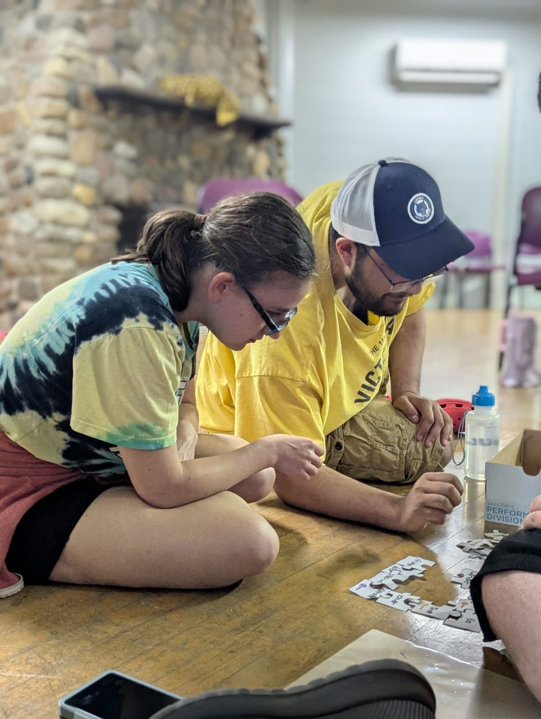 Woman and man on the floor working on a puzzle. Both are looking down, focused. Indoor setting.