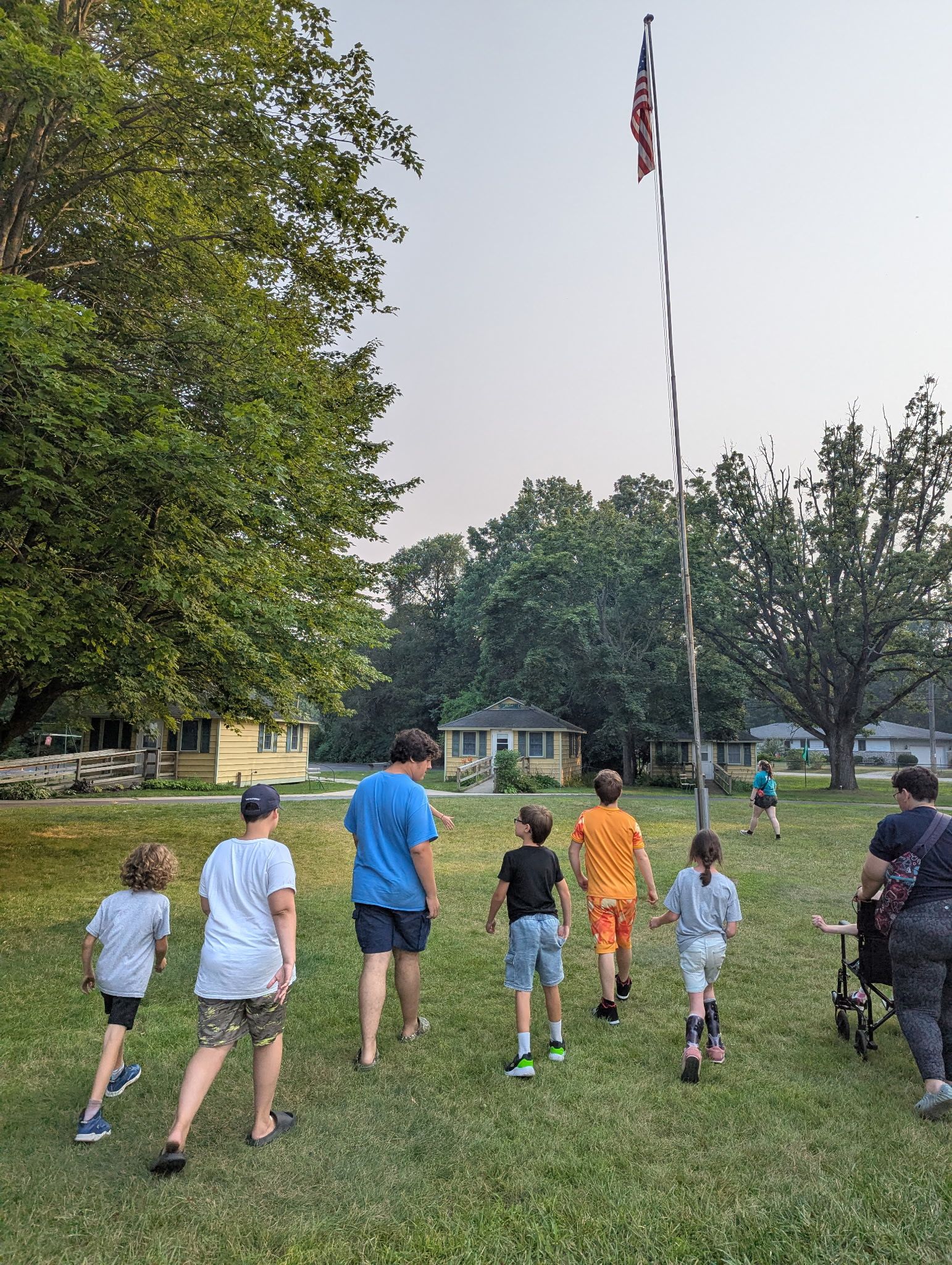 Group of people walking on grass towards a house, with a flag and trees in the background.