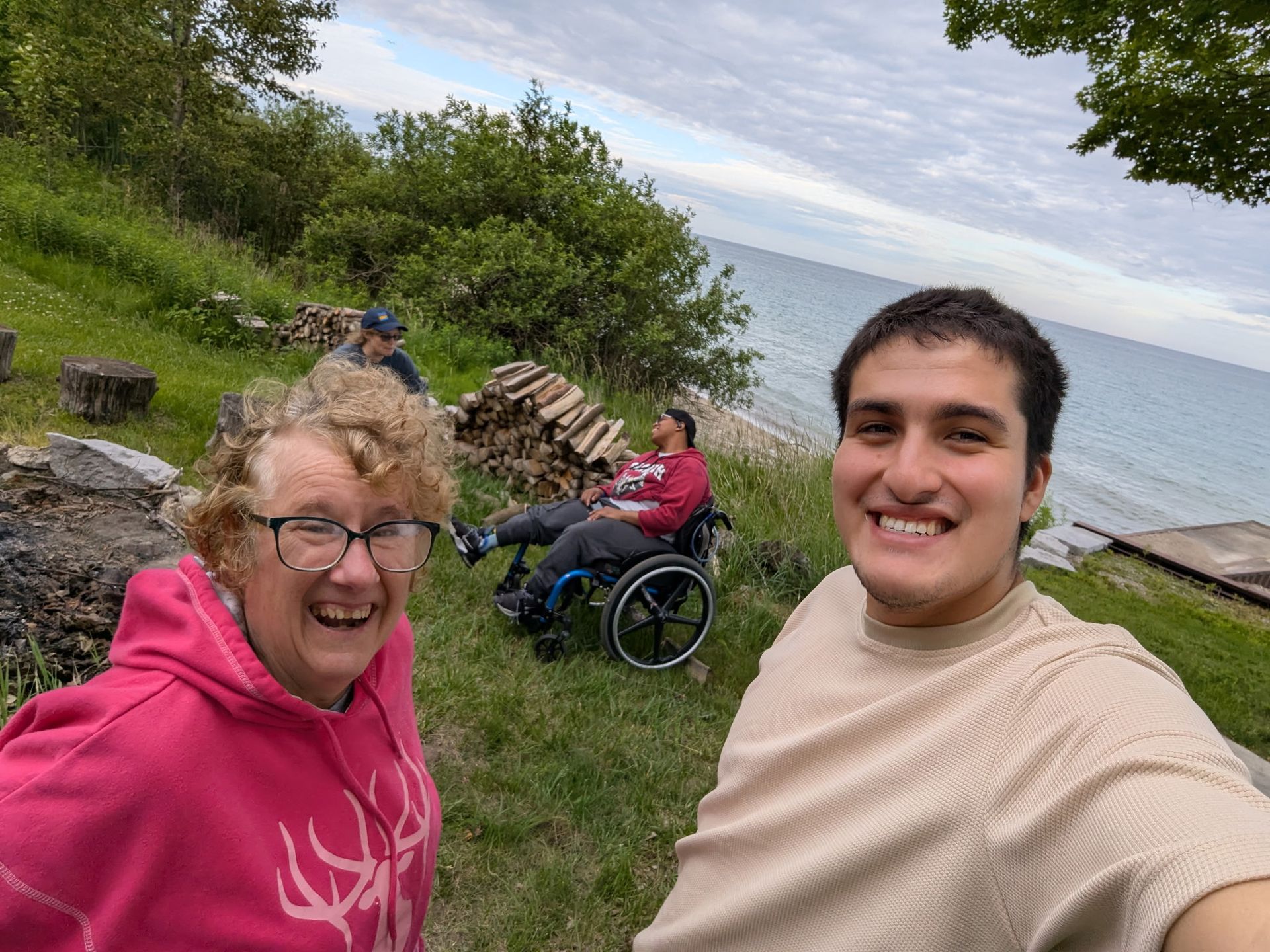 Group of people by a lake, including someone in a wheelchair. Smiling people and green grass.