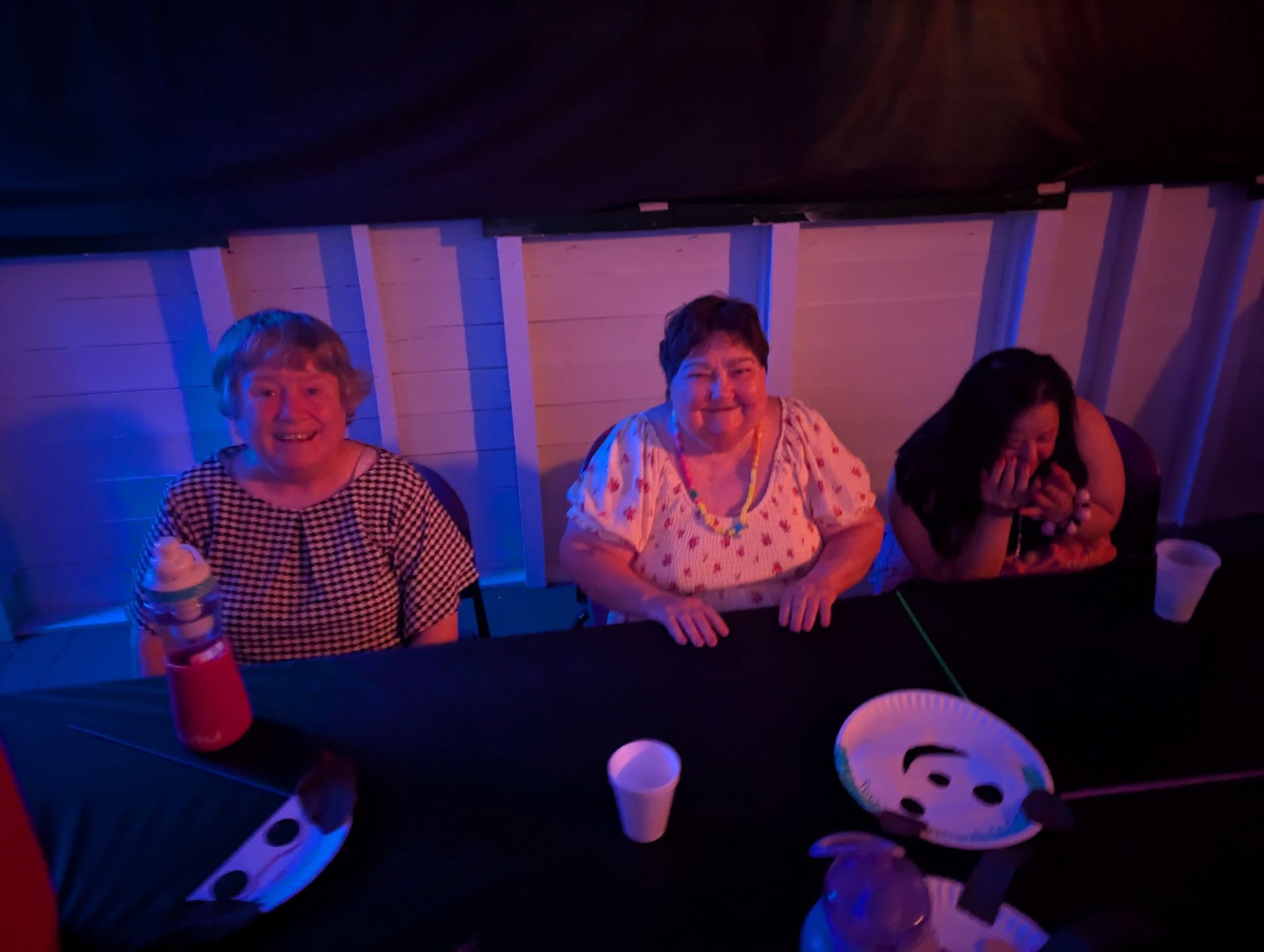 Three women seated at a black table under blue and red lights. Two are smiling, the third is covering their face.