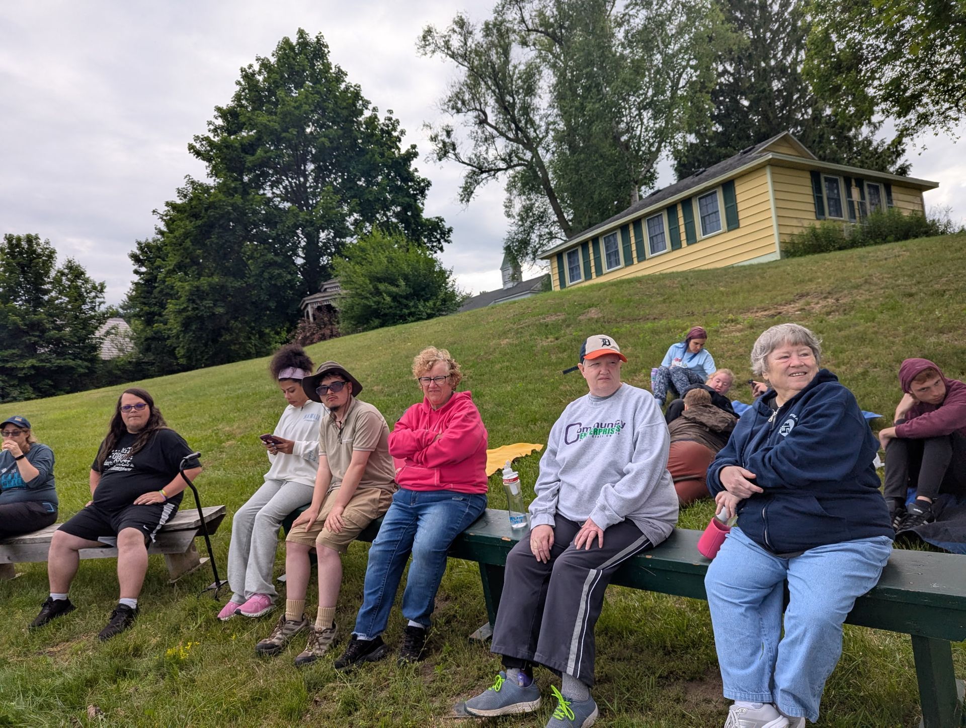 Group of people sit on a bench on a grassy hill, looking toward something out of frame, with a yellow building behind them.