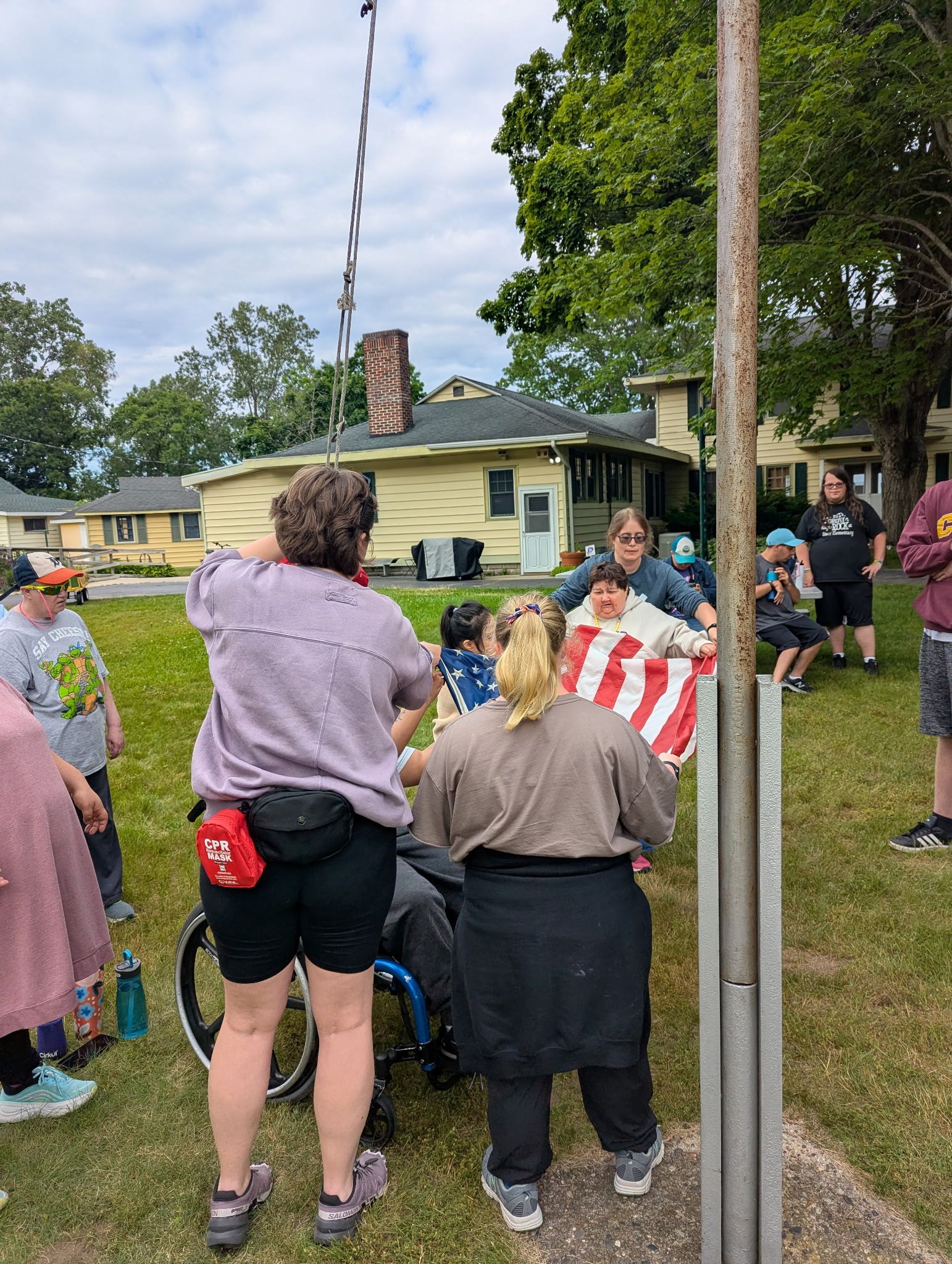 Group of people around a flagpole, one in a wheelchair. Outdoors, cloudy day, green grass, houses in the background.