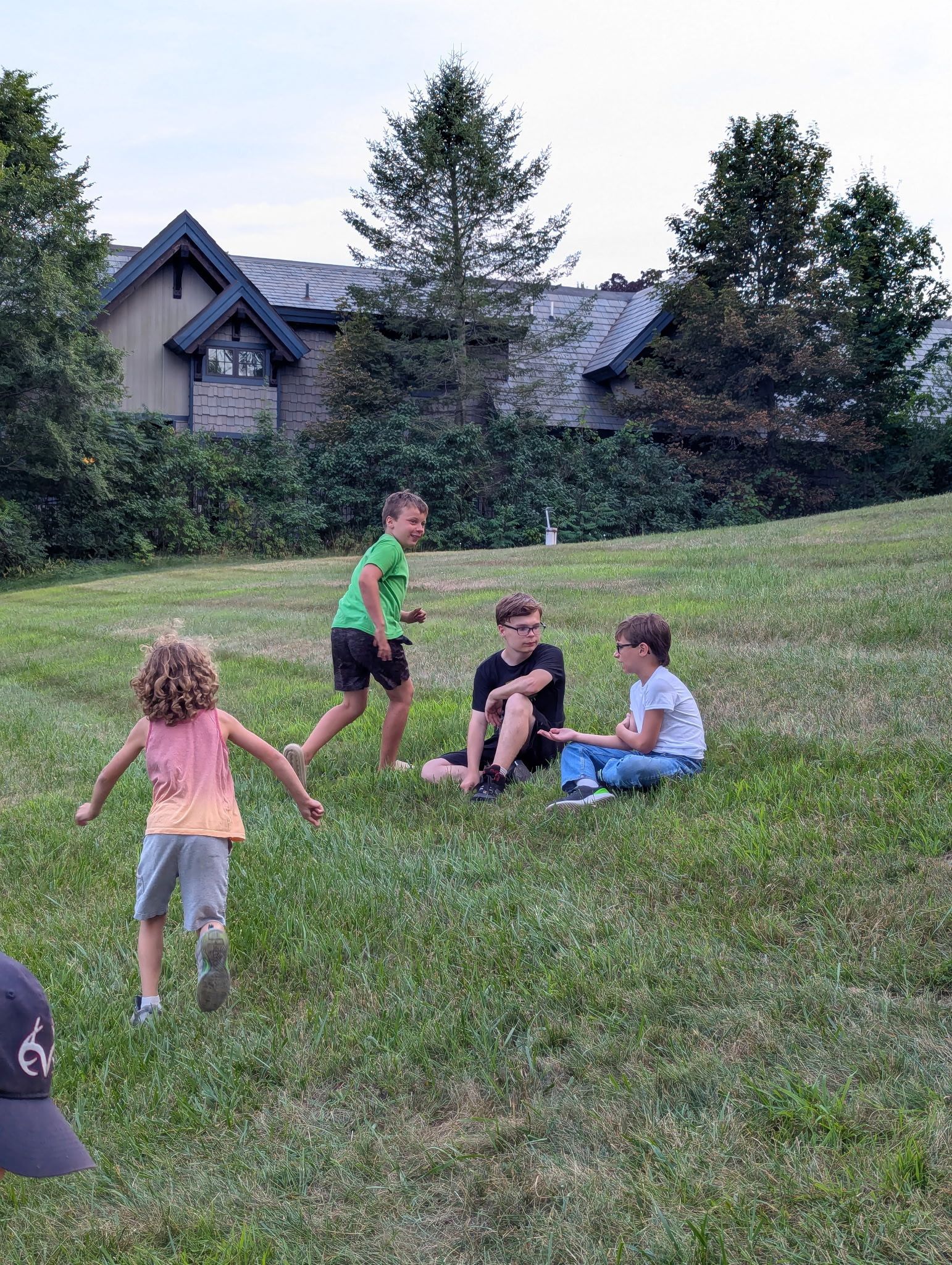 Children playing on a grassy hill. One runs, others sit and chat near a building.
