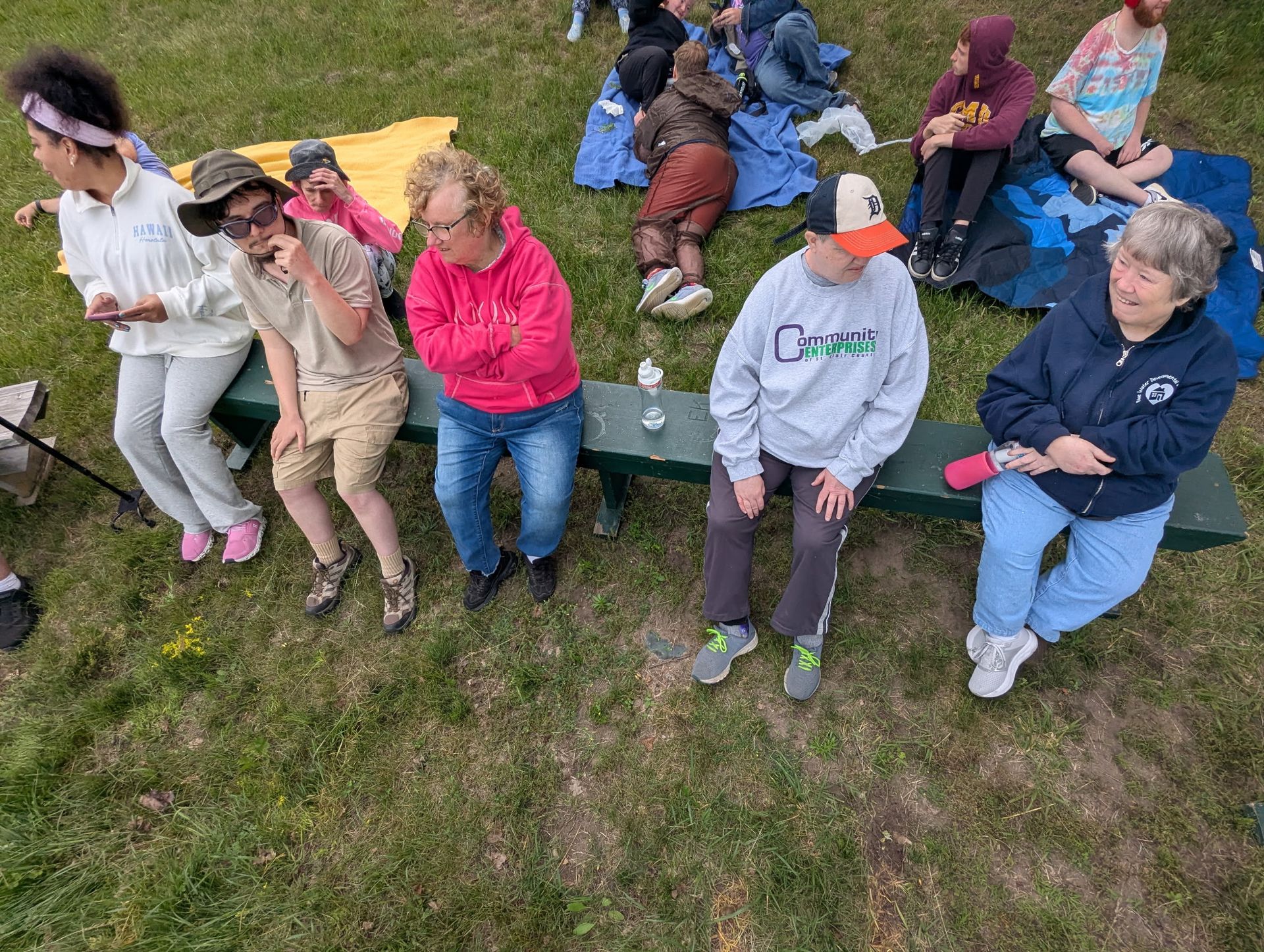 People seated on a green bench in a grassy area, some looking towards something.