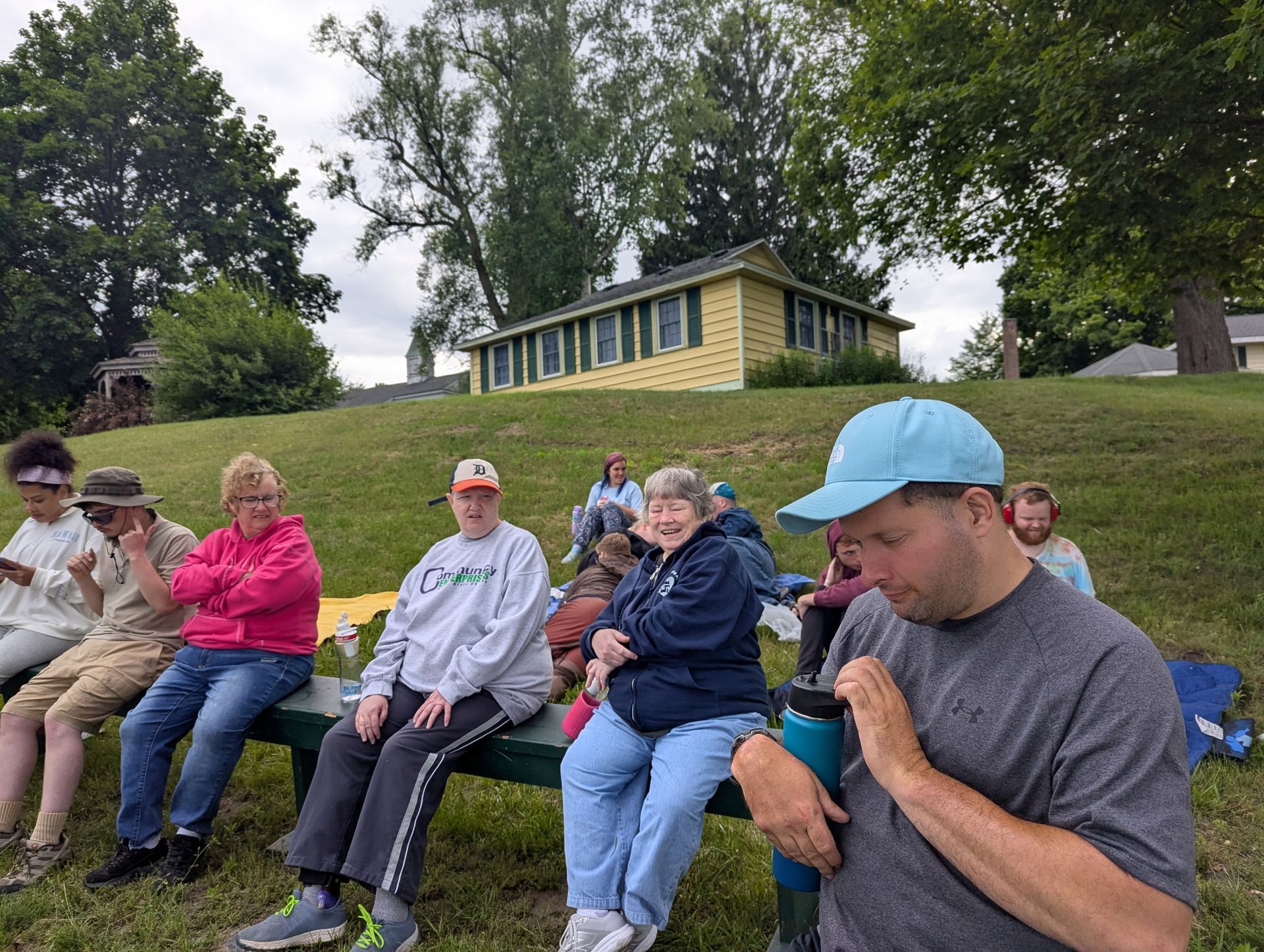 Group of people on a bench, looking towards the camera, in front of a yellow building on a grassy hill.