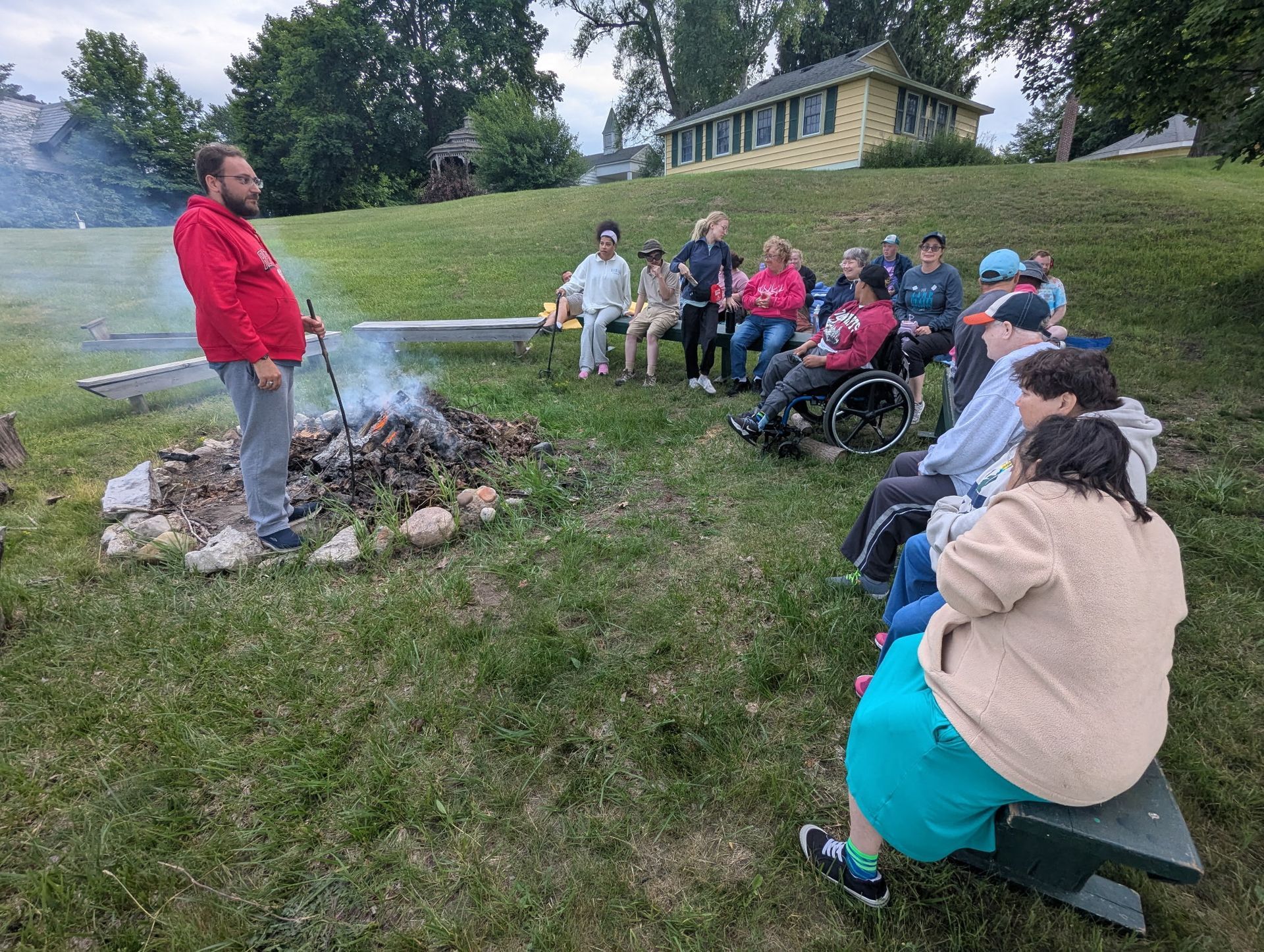 A group gathers around a campfire as a man in red talks to them in a grassy area.