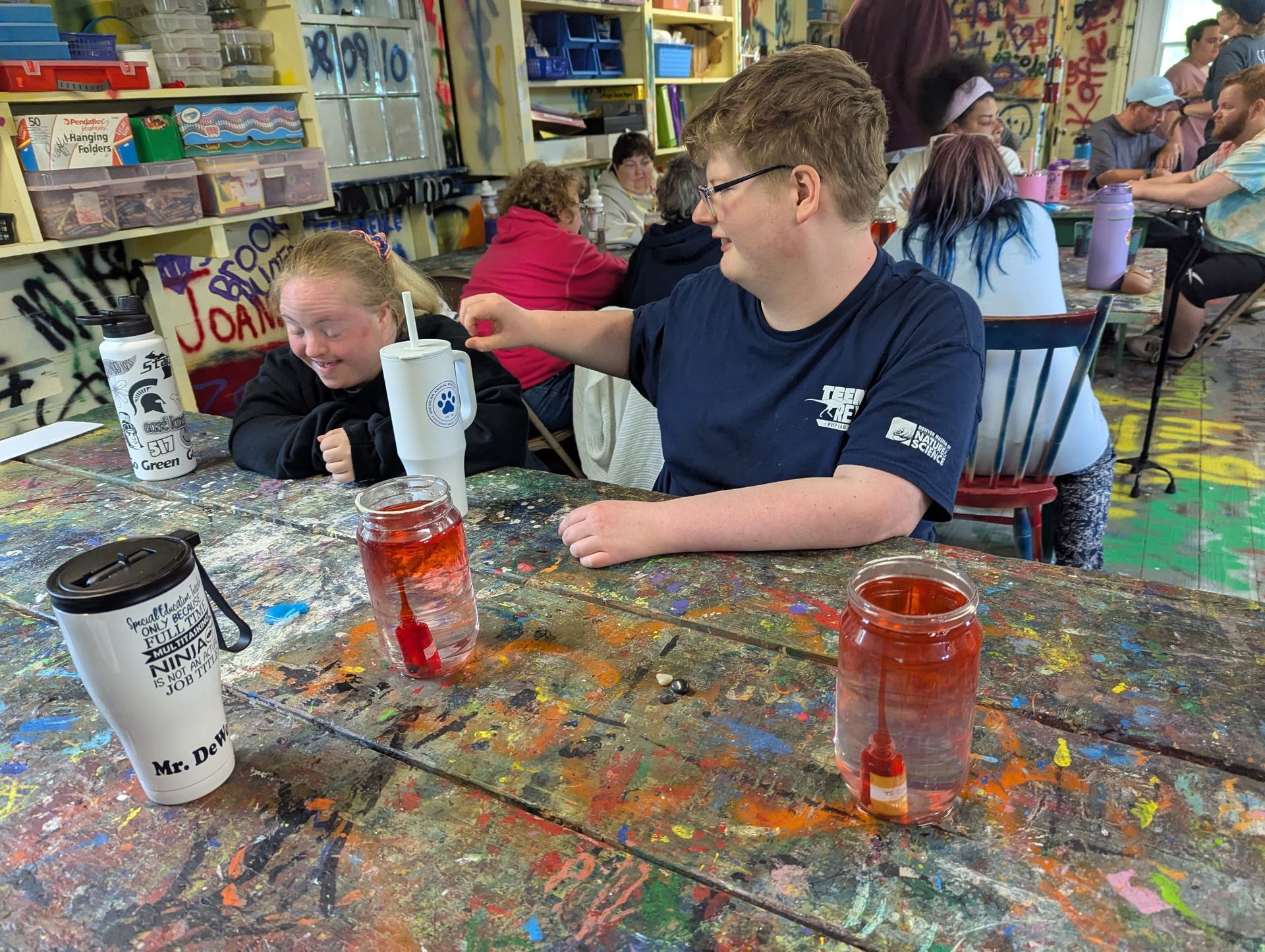 Two people at a colorful table, possibly art class. One points at a glass with red liquid. Others in background.