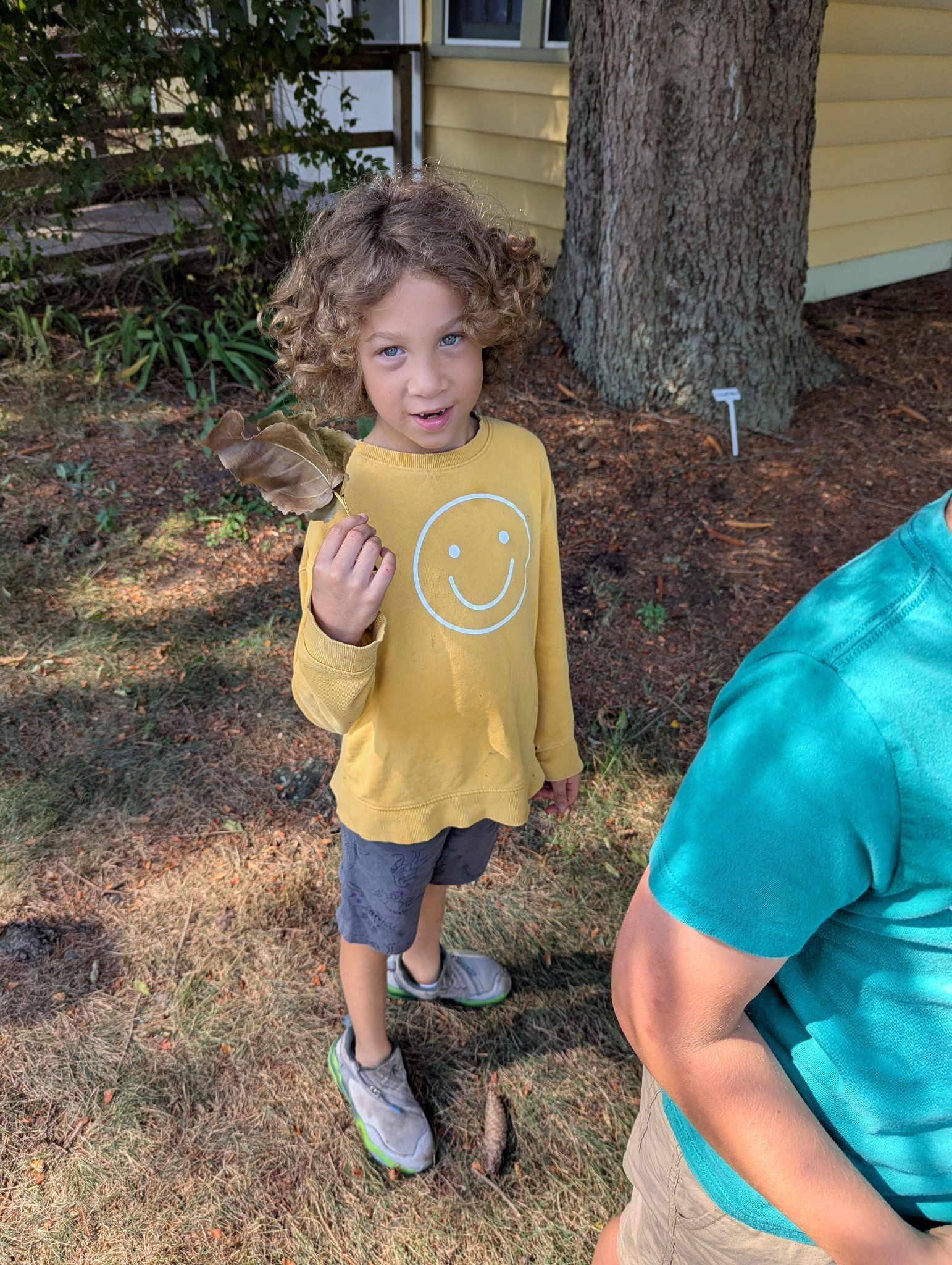 Boy in mustard shirt holding a leaf, smiling. Outdoor setting.