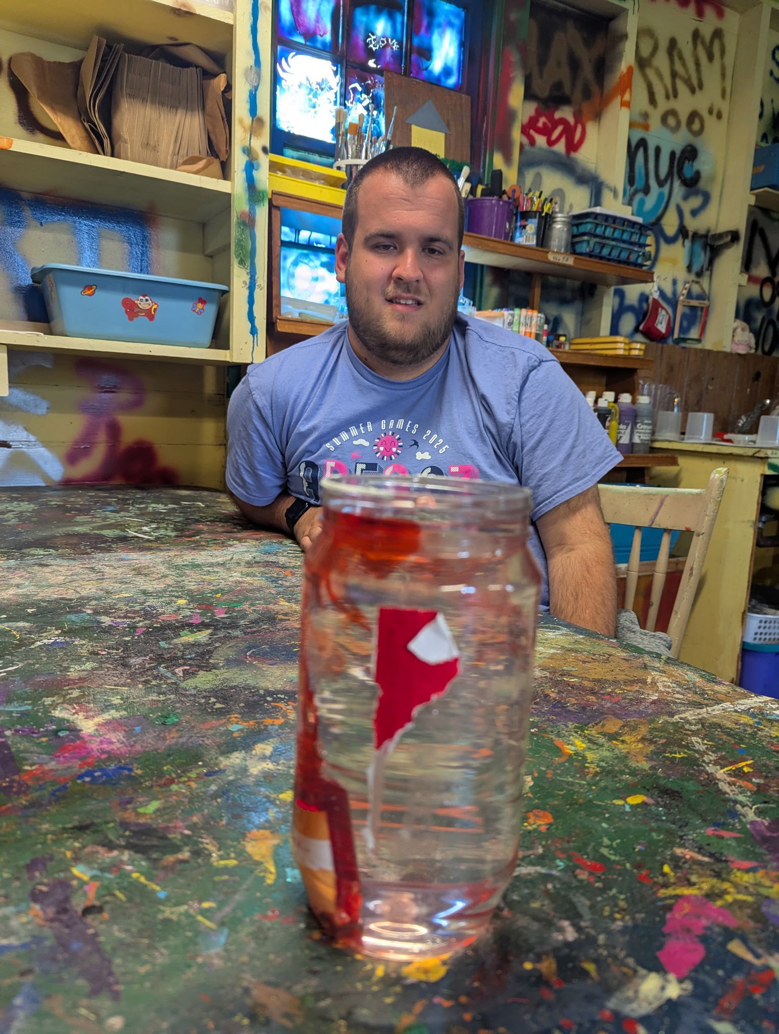 Man with disabilities smiles, behind a jar with red paper inside. Art studio setting, colorful, messy table.