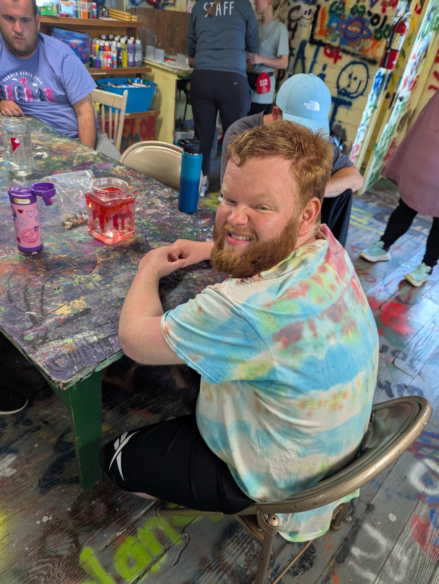 Man with red beard and tie-dye shirt smiles at the camera, sitting at a paint-splattered table in a studio.