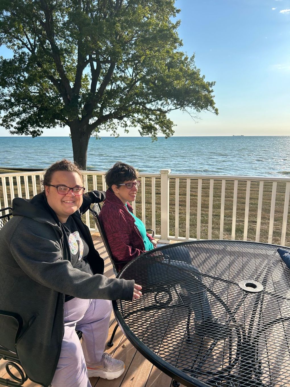 Two people seated at a patio table overlooking a lake. The person on the left smiles, and both are outdoors.