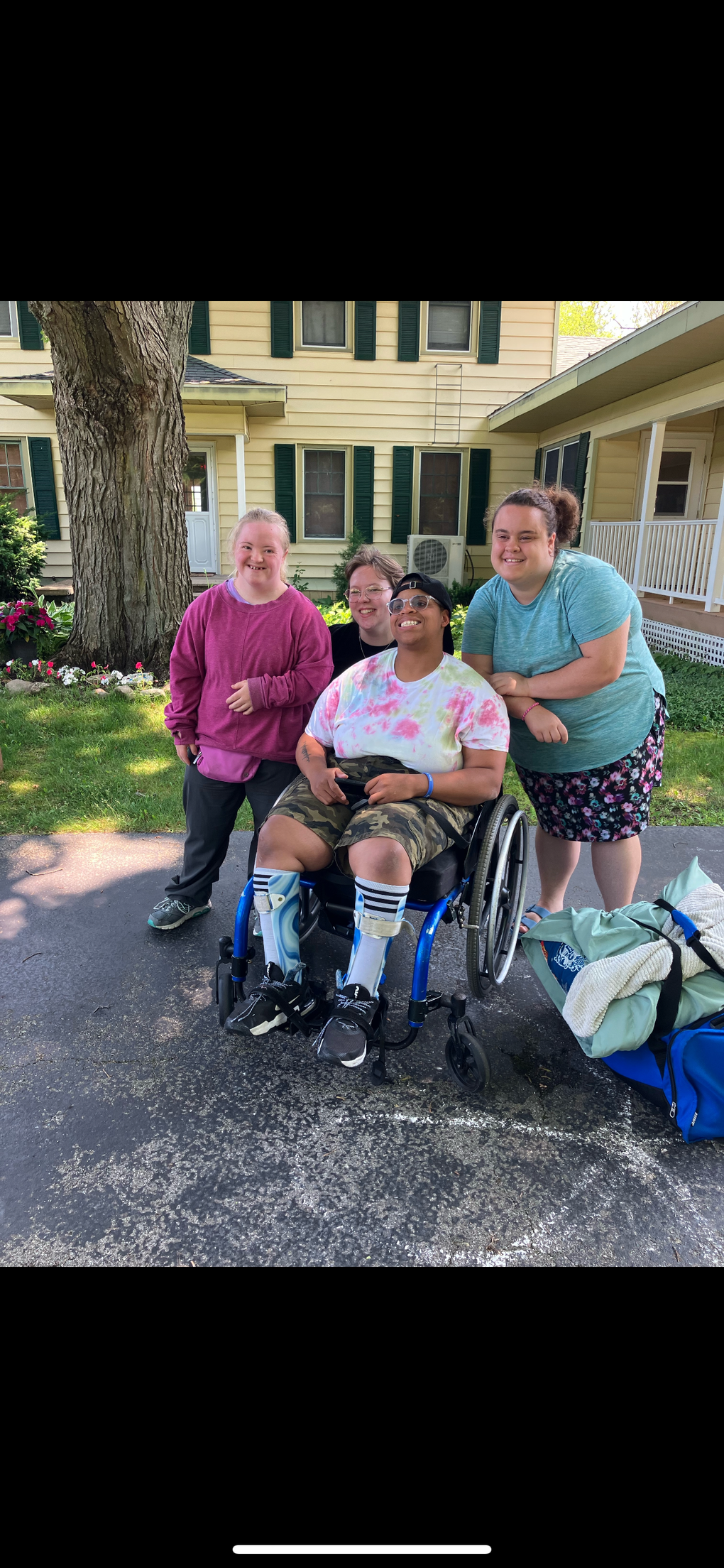 Four people pose outdoors: one in a wheelchair, others standing. A yellow house in the background.