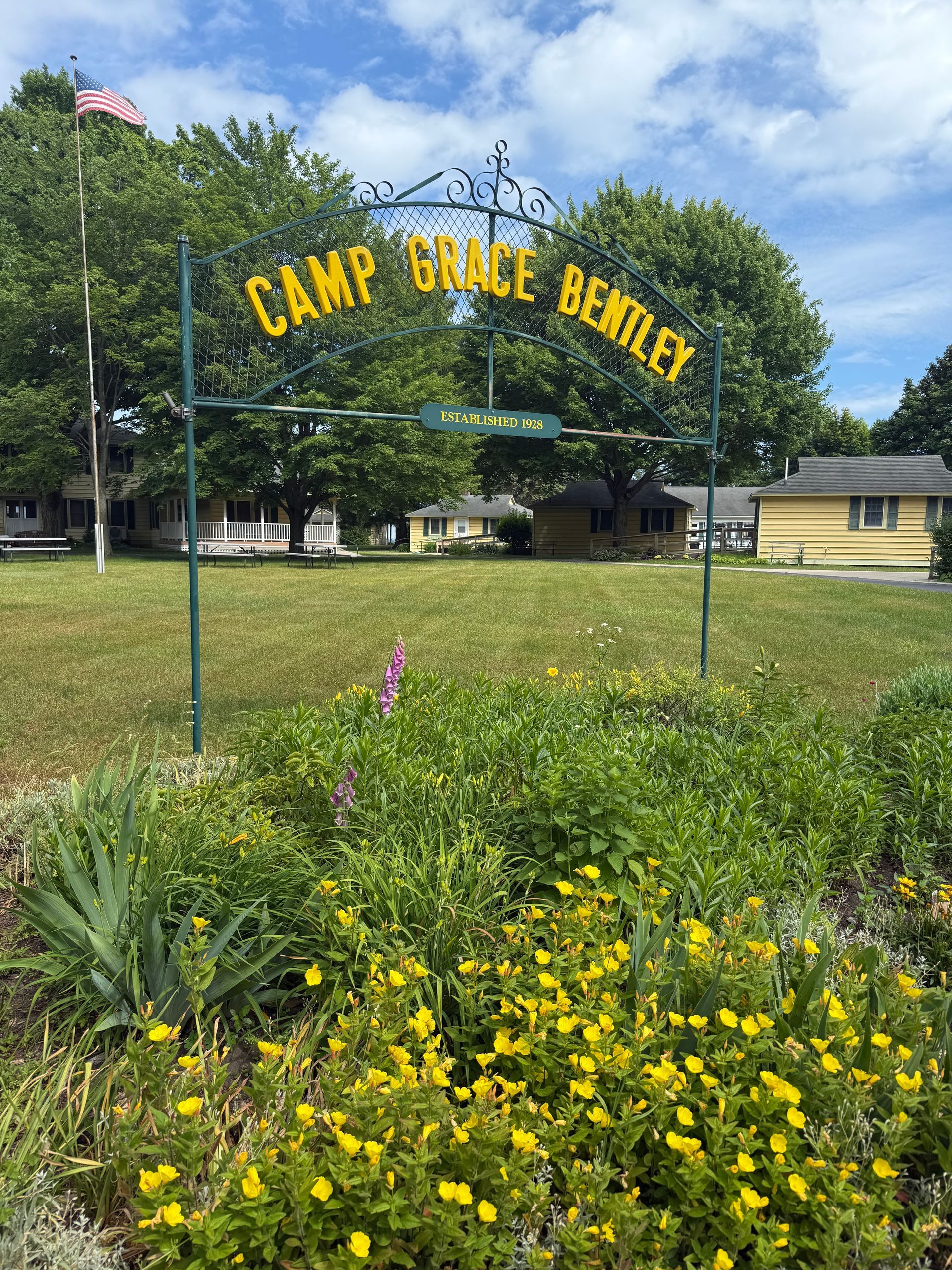 Sign for Camp Grace Bentley over a grassy field with buildings and flowers.