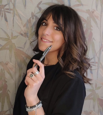 Donna con i capelli scuri, con una penna in mano, con aria pensierosa e gioielli, seduta contro una parete decorata.